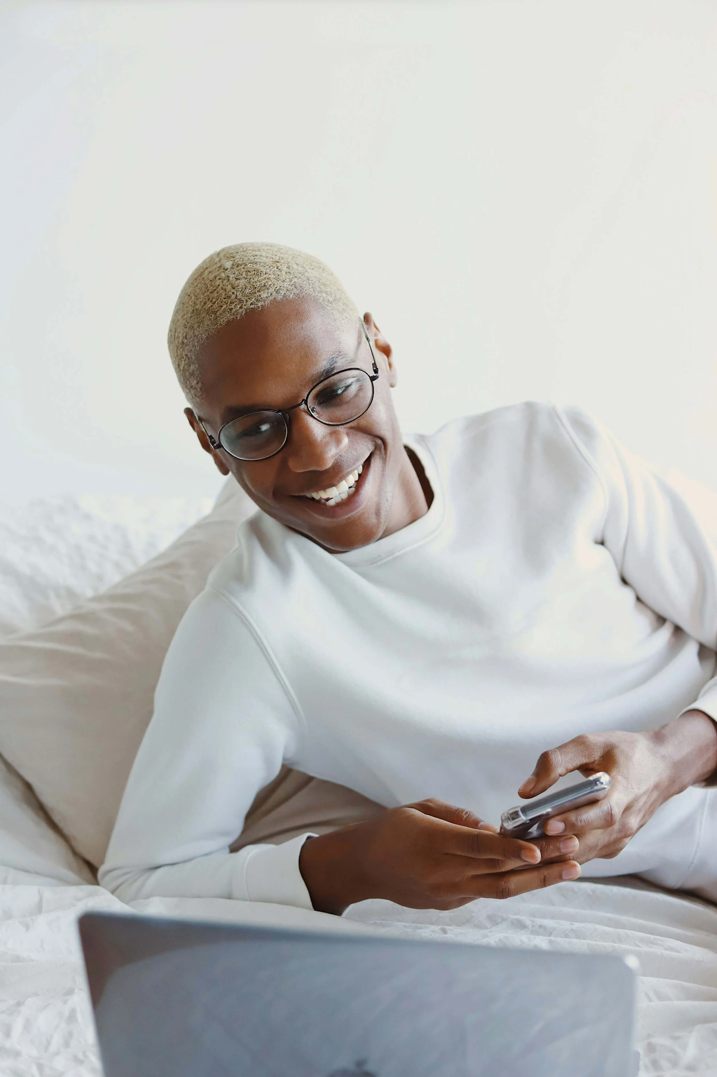 A young woman with short blonde hair and glasses lying on a bed, smiling while looking at her smartphone with a laptop in front of her.