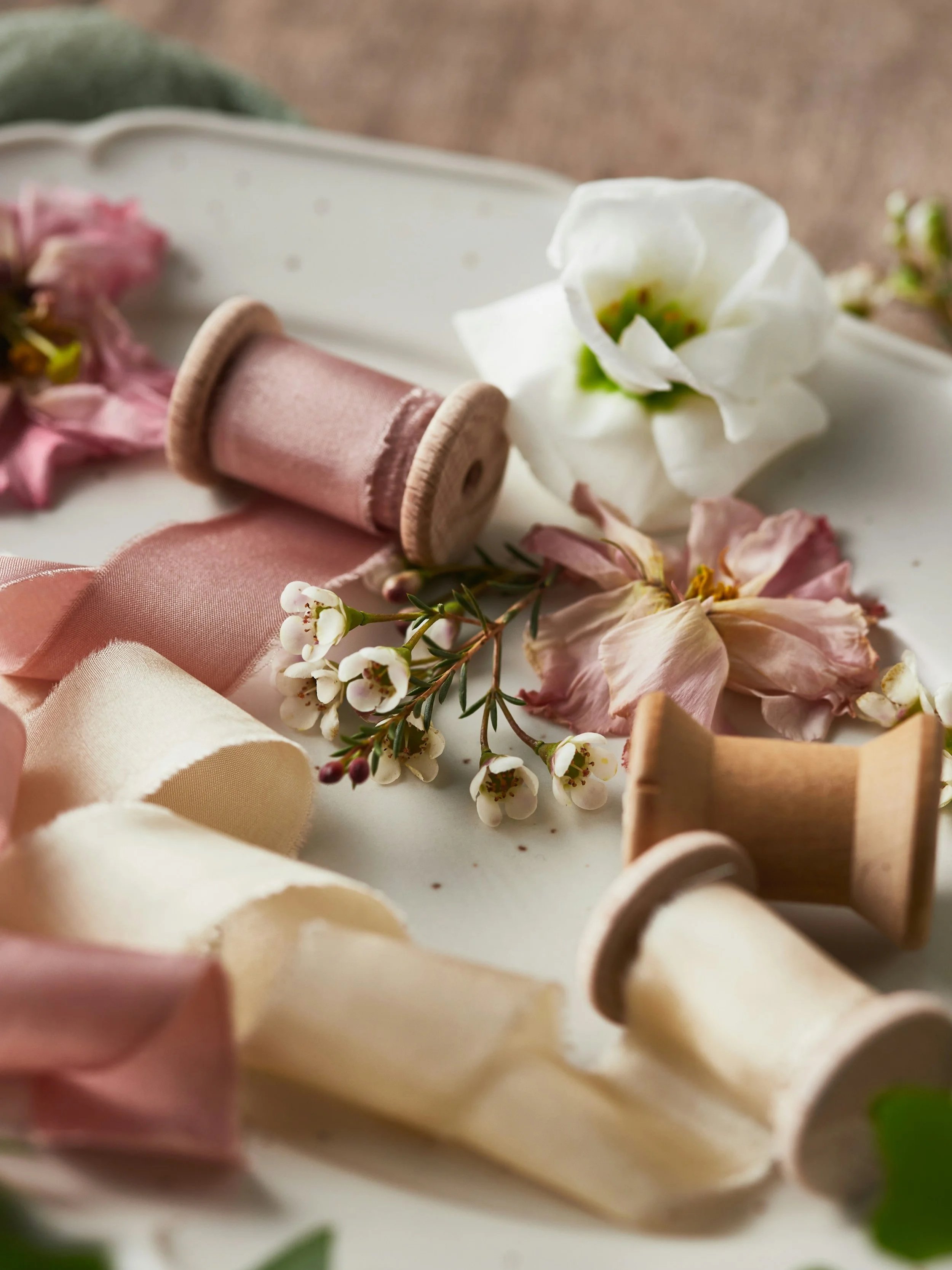 Soft pastel-colored fabric ribbons, wooden spools, and delicate flowers arranged on a white surface.