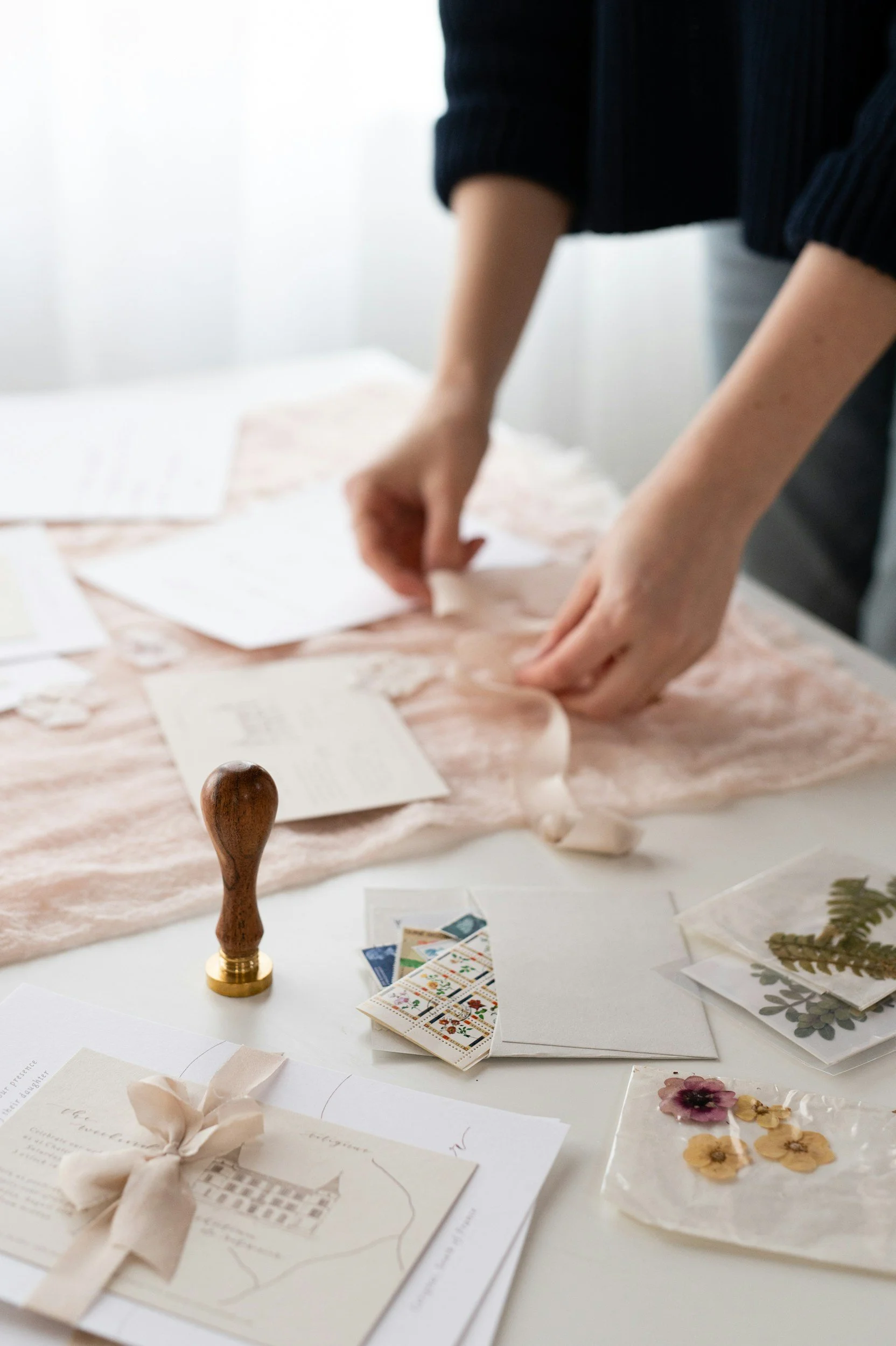 Person's hands creating a wedding invitation or craft with various papers, stickers, and embellishments on a white table.