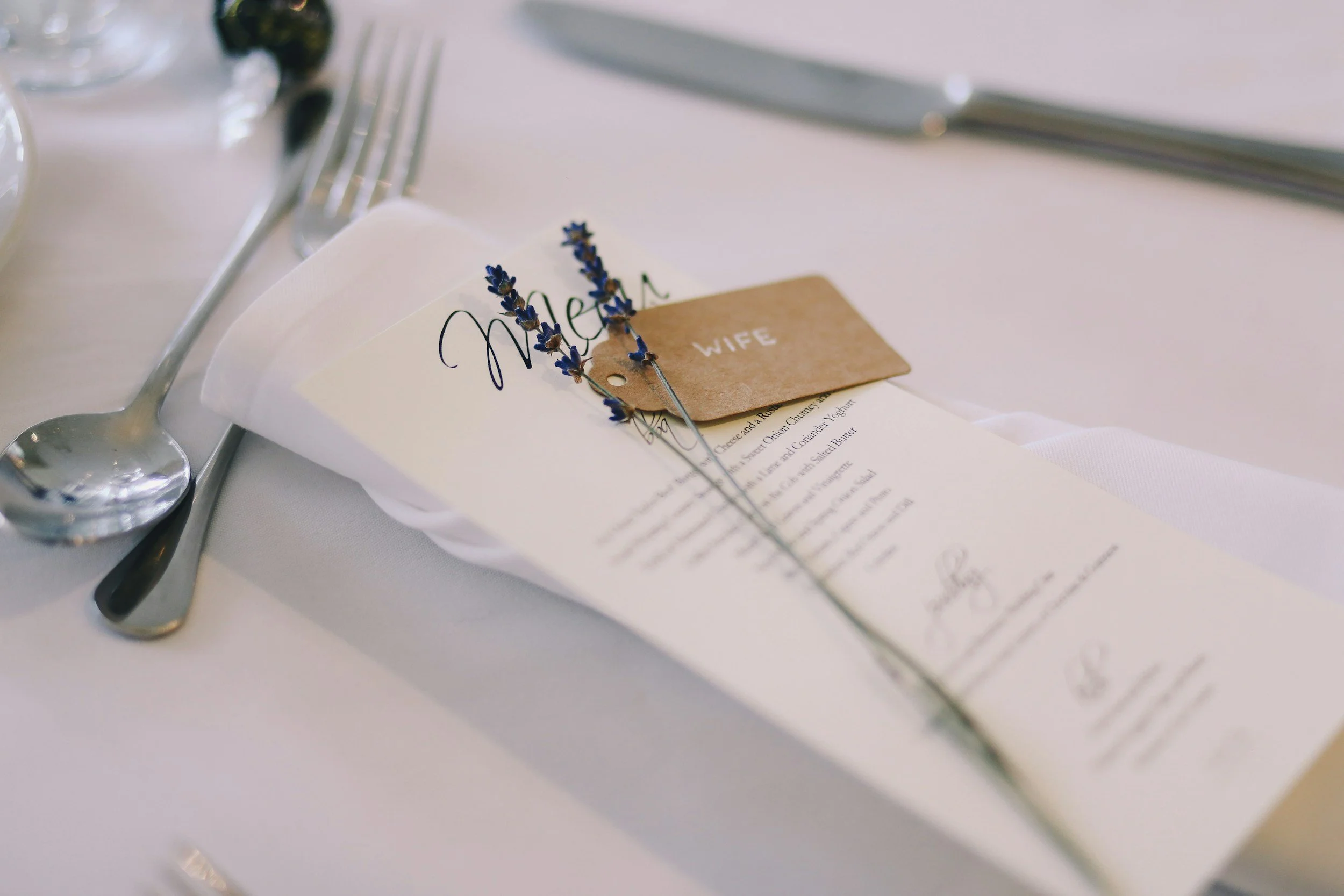 A place setting at a wedding reception with a menu, a lavender sprig, and a brown place card labeled "WIFE" on a white tablecloth.