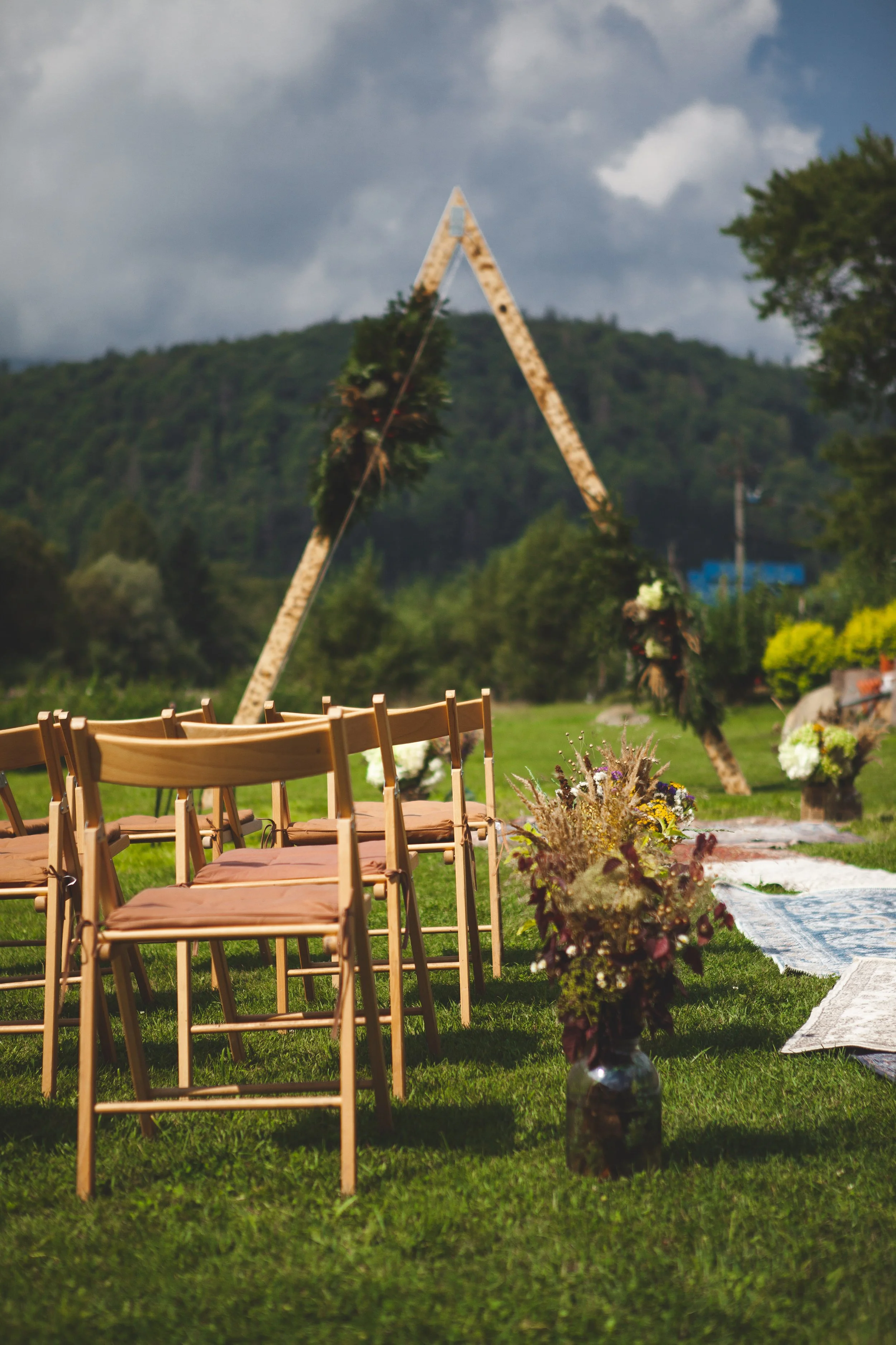 Outdoor wedding setup with wooden chairs arranged on grass, floral arrangements, and a triangular arch decorated with greenery, set against a scenic mountain backdrop under cloudy sky.
