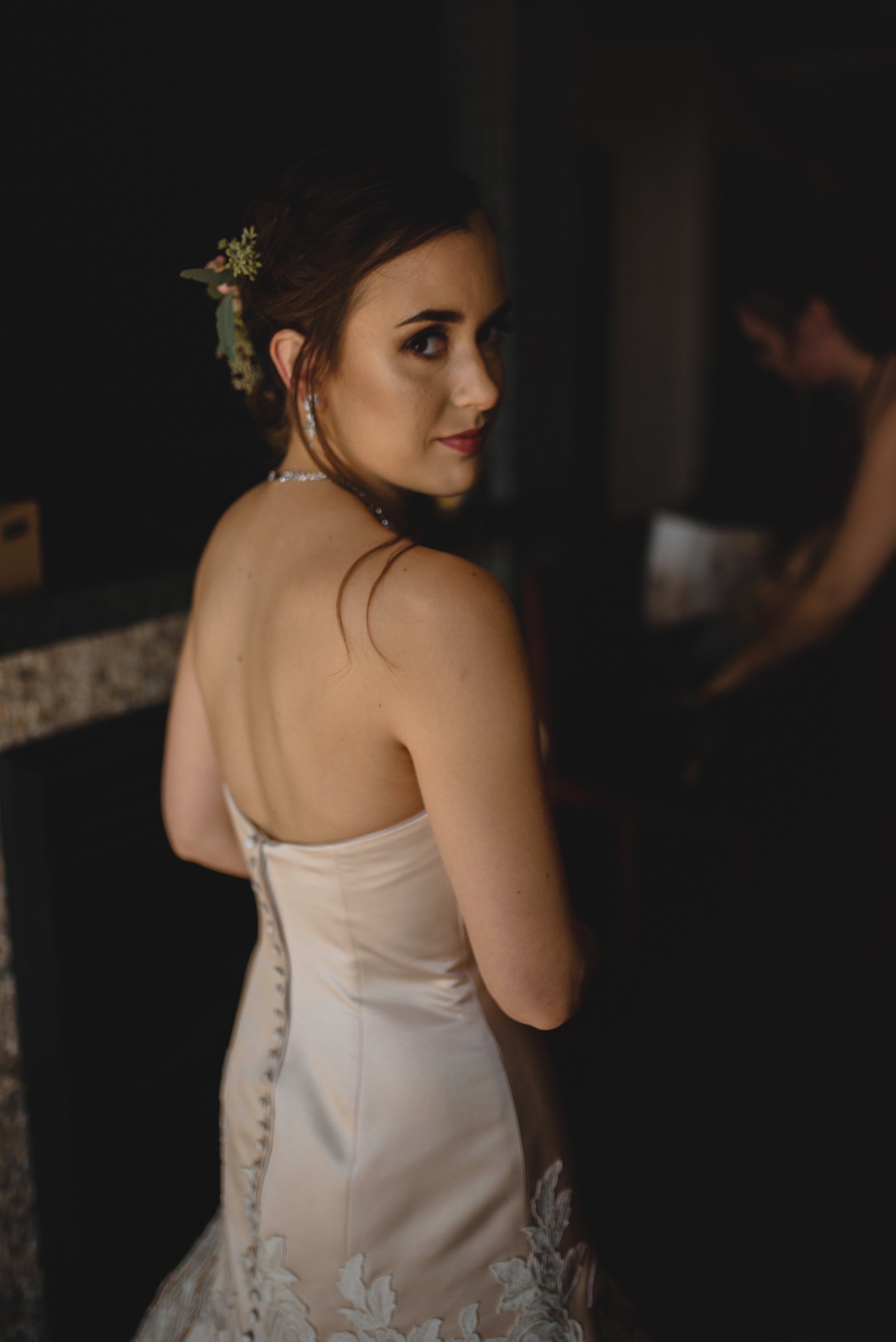 A woman in a wedding dress looking over her shoulder, with a dark background and another person in the background.