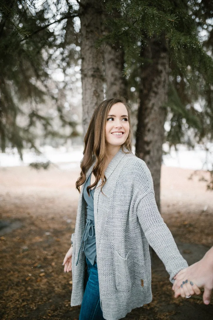 A woman with long brown hair smiling and holding hands with someone outdoors under a tree.