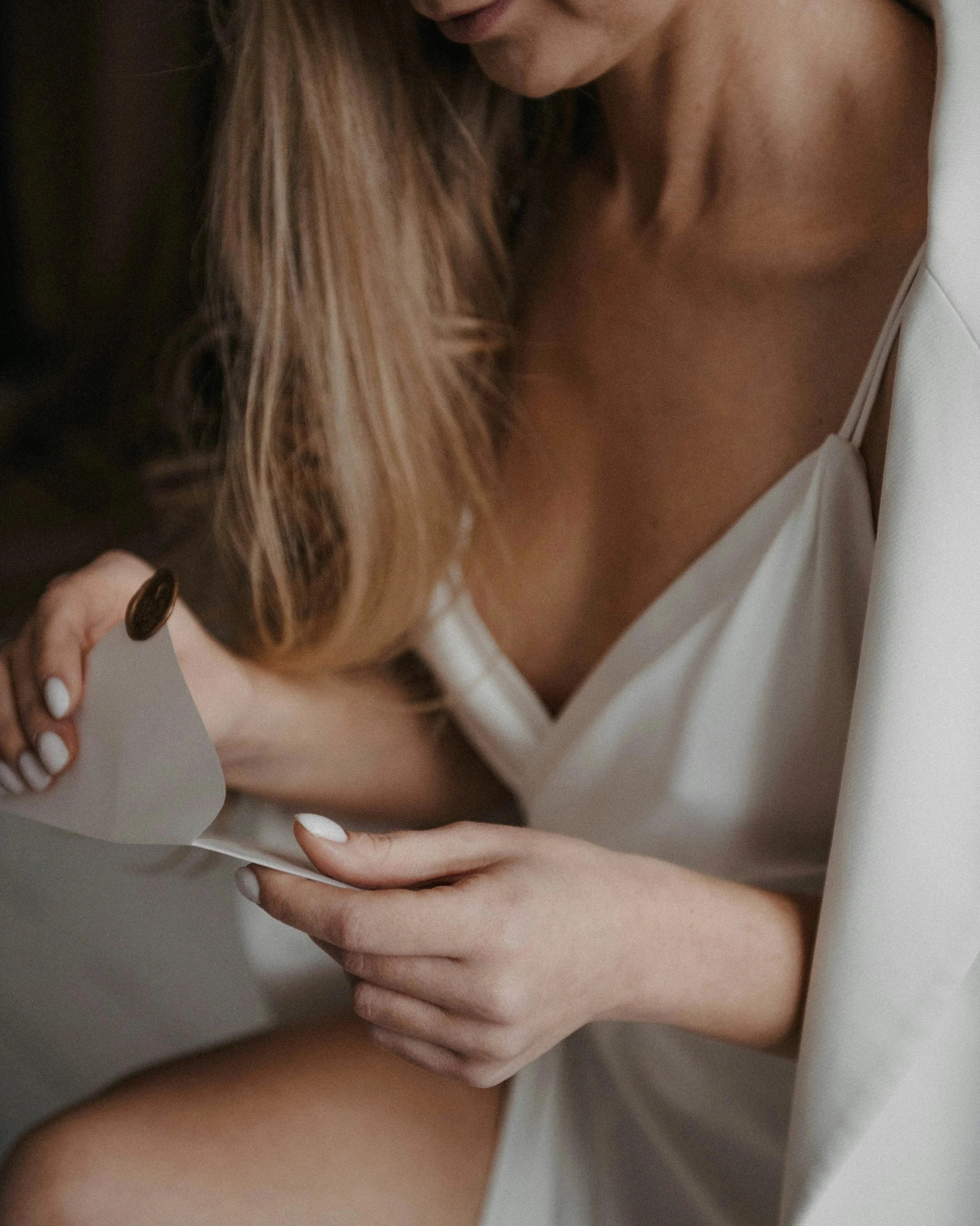 A woman with long, wavy hair wearing a white satin slip dress, seated and reading a letter with a gold coin attached to it.