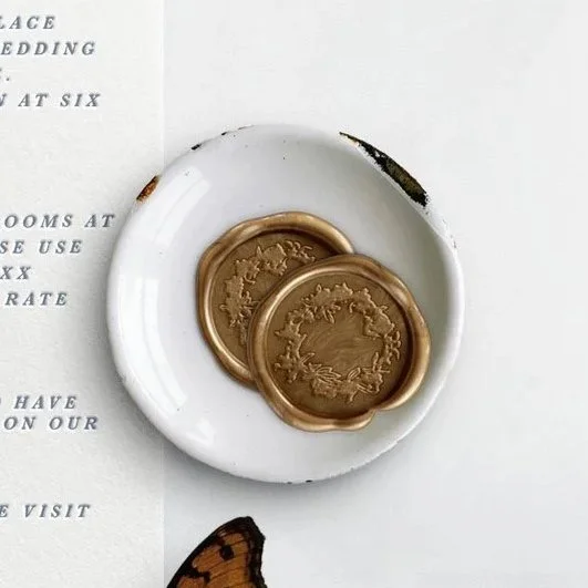 Two gold wax seals on a white plate, partially overlapping, on a white surface next to a piece of paper and a butterfly wing.