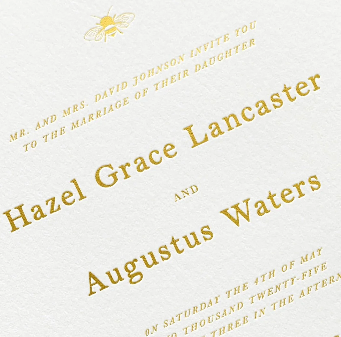 Close-up of a wedding invitation with gold foil text and an embossed bee illustration.