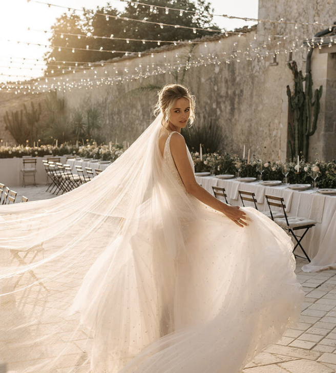 A bride in a white wedding gown with a long veil outdoors, with string lights and tables set for a celebration in the background.