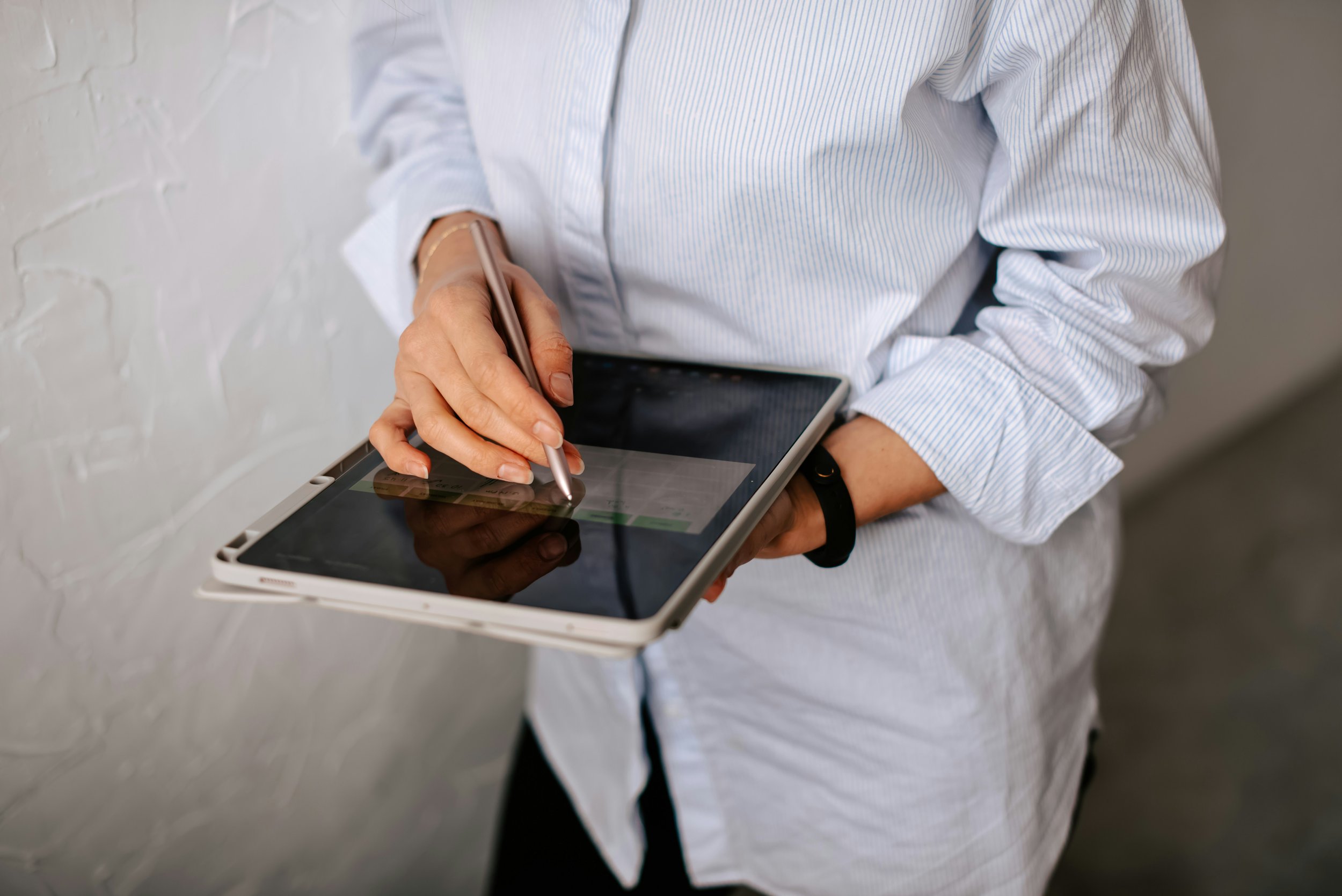 Person holding a tablet and using a stylus to write or draw on the screen, wearing a light blue and white striped shirt, against a white textured wall.