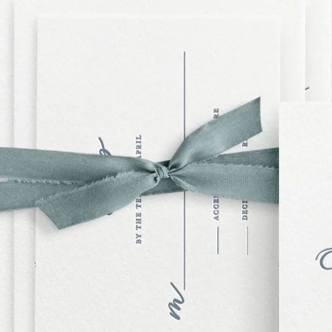 Close-up of a white wedding invitation with gray ribbon tied in a bow, partially visible text, and lines for date, time, and location.