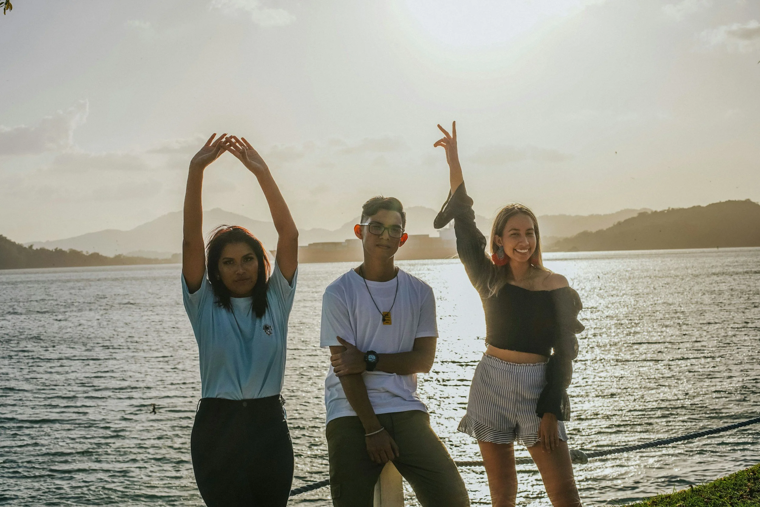 Four young people standing in front of a body of water during sunset, with mountains in the background. They are posing and smiling, with some raising their hands.