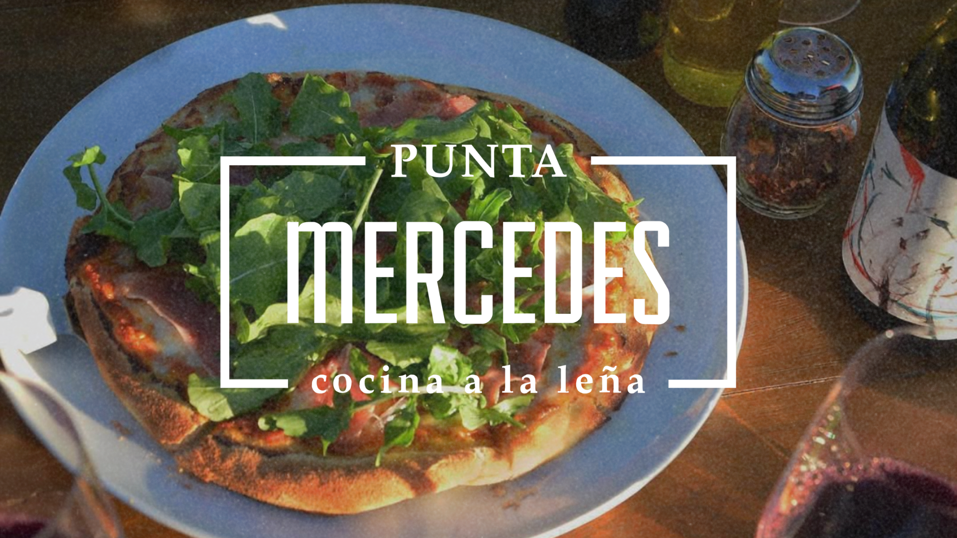 Wood-fired pizza topped with arugula on a plate, surrounded by a wine glass, olive oil bottle, and spice shaker; with "Punta Mercedes" text overlay.