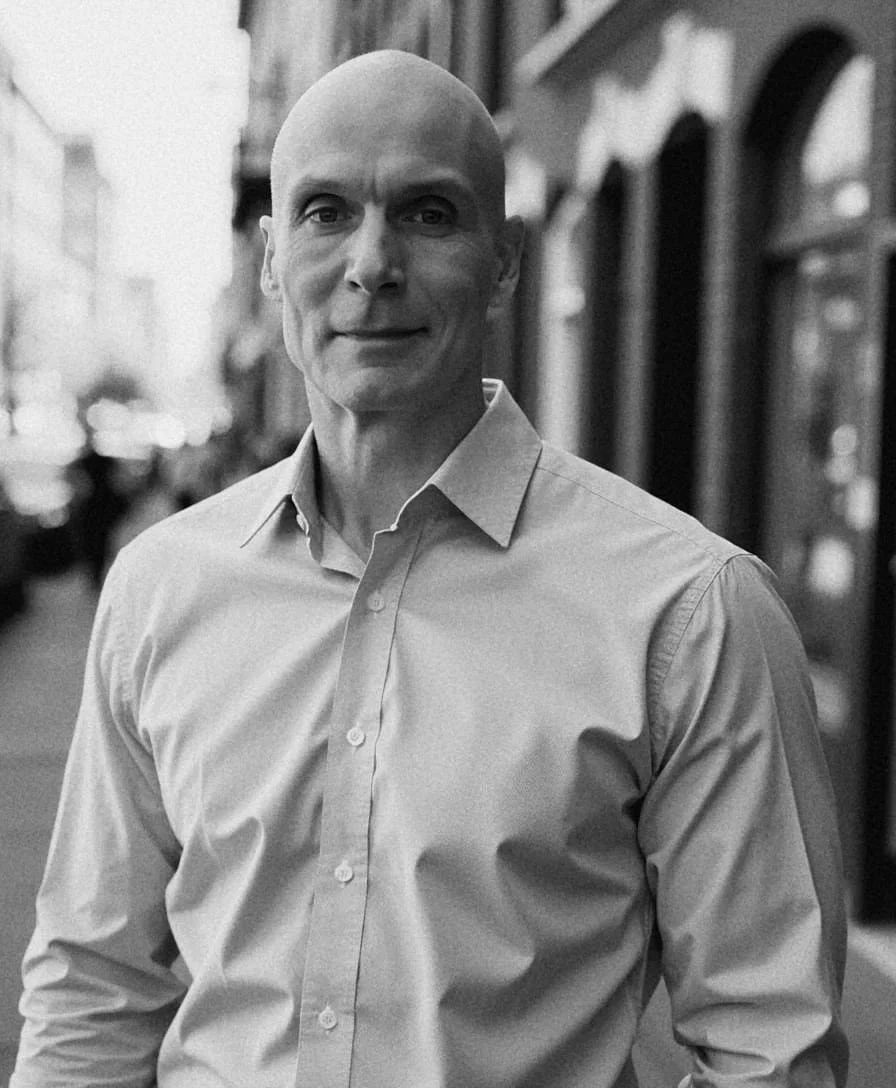 Black and white photo of Chris O'Gorman in a button-up shirt, standing on a street with buildings in the background.