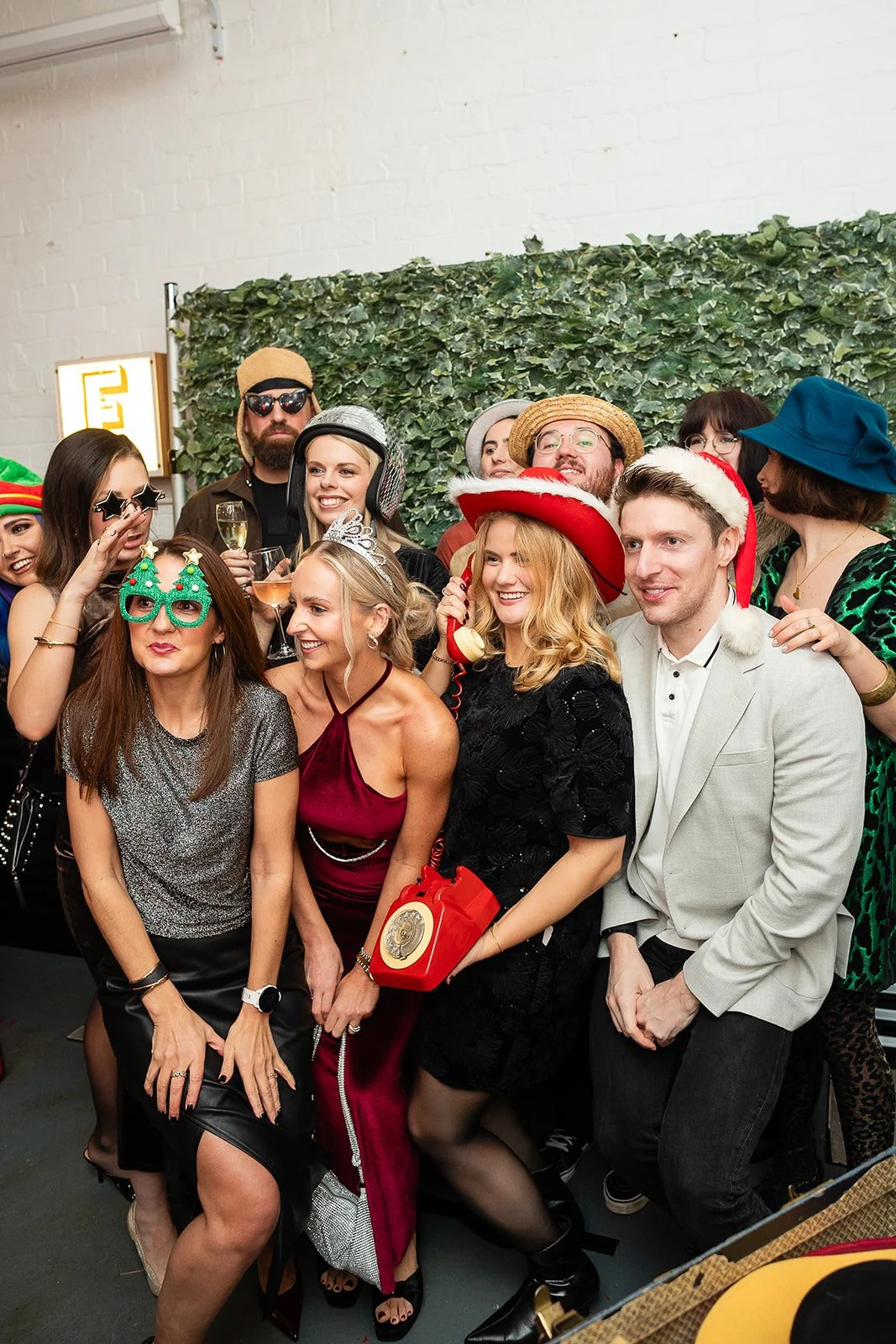 Group of people celebrating at a party, wearing festive hats and glasses, some holding drinks, with a green leafy backdrop.