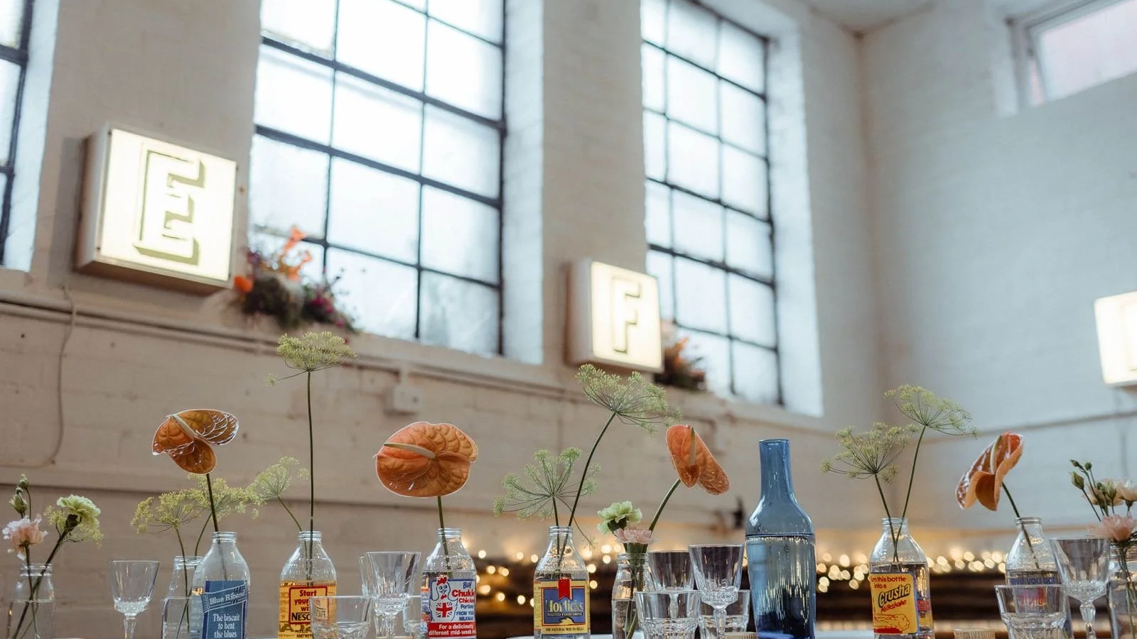 A table decorated with various vases holding flowers and foliage, set in front of large industrial-style windows and illuminated walls in a bright, airy room.