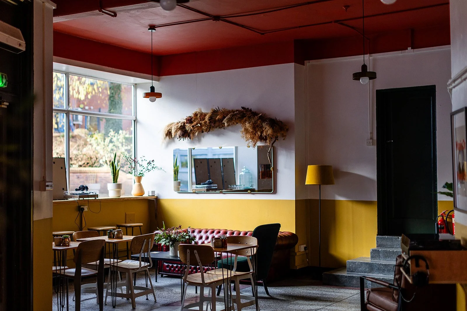 Front Room at The Hide wedding and events venue, Sheffield. Images shows wooden tables and chairs with a red Chesterfield in the background. Walls are paint mustard at the bottom and then white at the top. There is large mirror on the wall with a dri