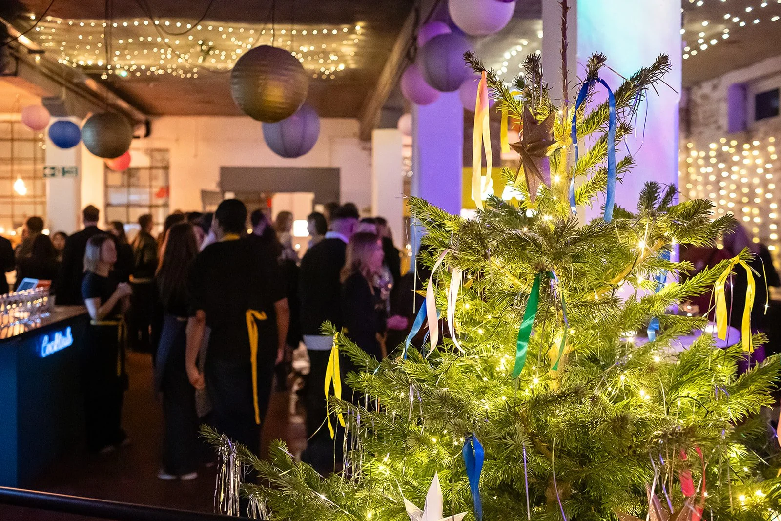 A decorated Christmas tree with colorful ribbons and lights in the foreground, with a busy party scene of people socializing and hanging out in a decorated venue with string lights and paper lanterns hanging from the ceiling in the background.