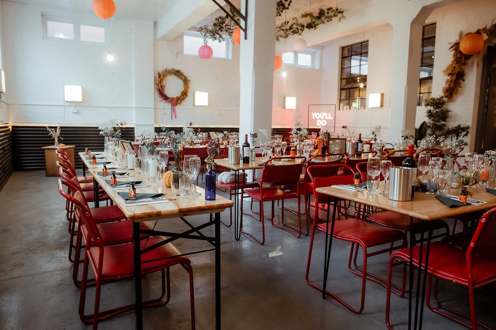 Warehouse space at The Hide, wedding and events venue, Sheffield. Images shows tables lined and dressed for a wedding using dried flowers and candles. Table are wooden with black legs and chairs are original cox chairs in red. Walls in the warehouse 