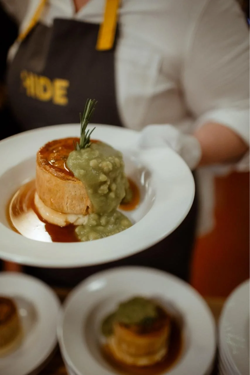 A plated gourmet dish featuring a homemade pie, topped with a sprig of rosemary, accompanied by mushy pies and brown gravy, served on white dishware.