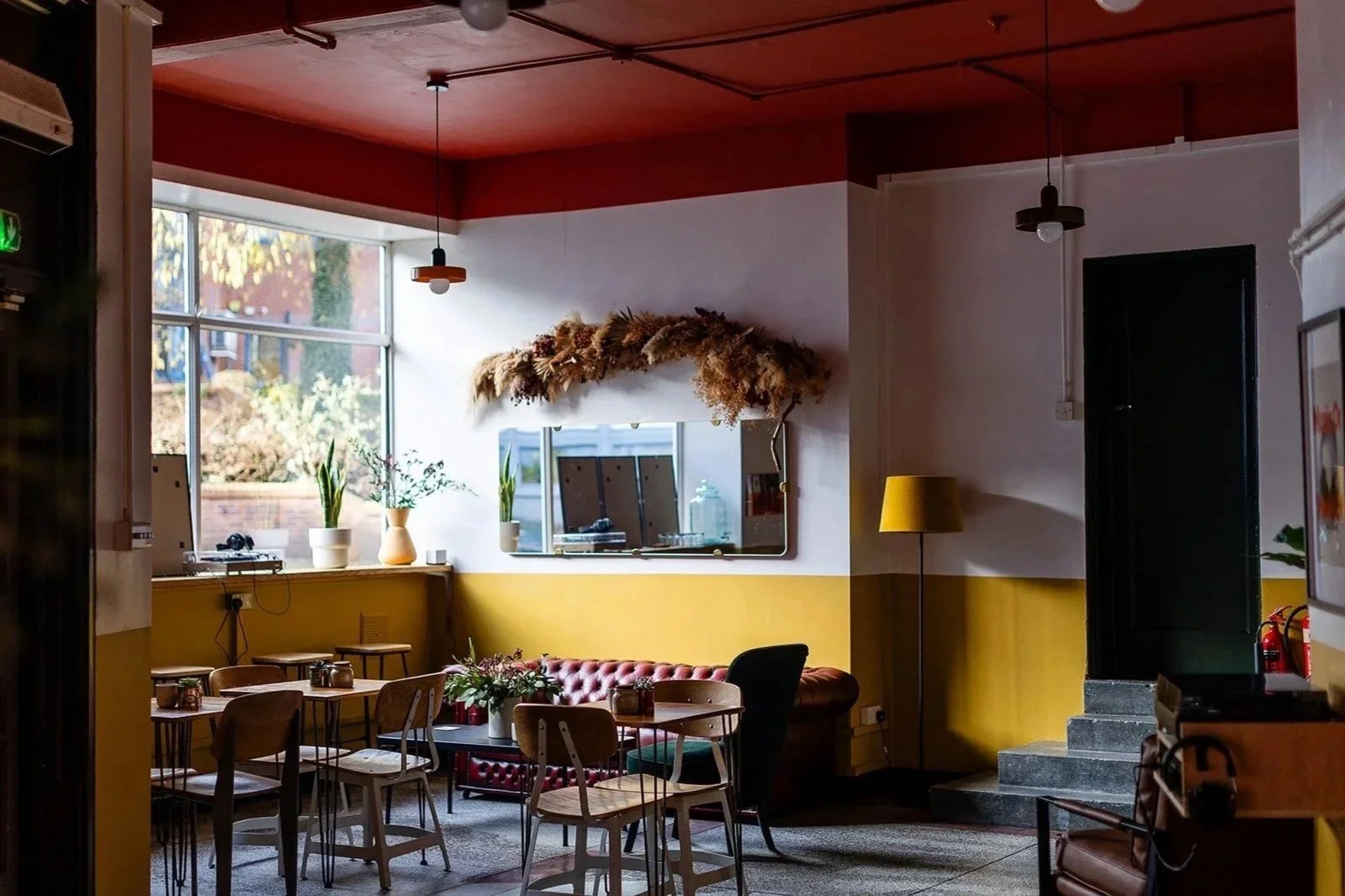 Front room coffee shop at The Hide, Sheffield. Walls are painted mustard half way and white at the top. There are plywood tables and chairs, and a red Chesterfield sofa in the background with a black coffee table. A large mirror hangs above the sofa.