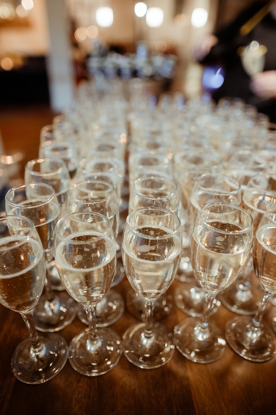 Multiple glasses of champagne on a wooden table in a blurry, warmly lit room.
