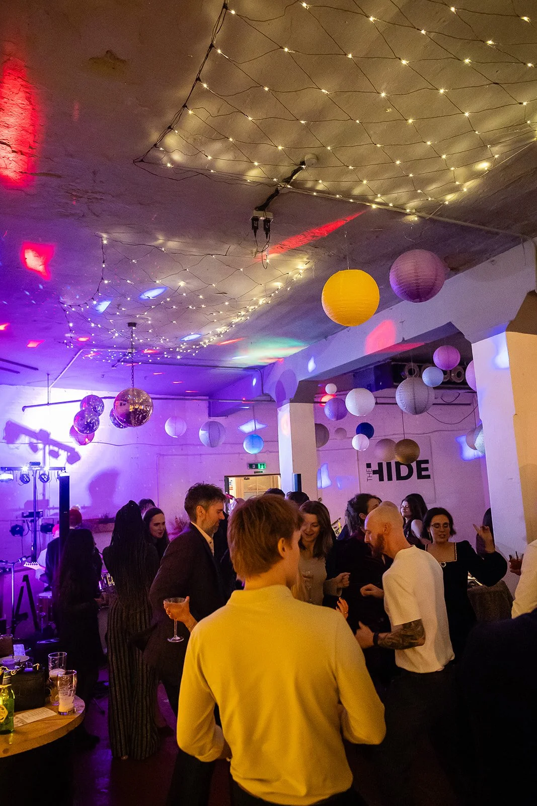 People socializing at a party with colorful lanterns and string lights hanging from the ceiling, in a decorated indoor space.