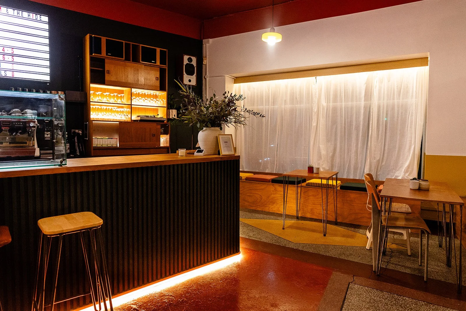 Interior of a modern cafe with a wooden counter, tables, chairs, and a cozy seating area with cushions. Soft lighting and a white curtain cover the back wall.
