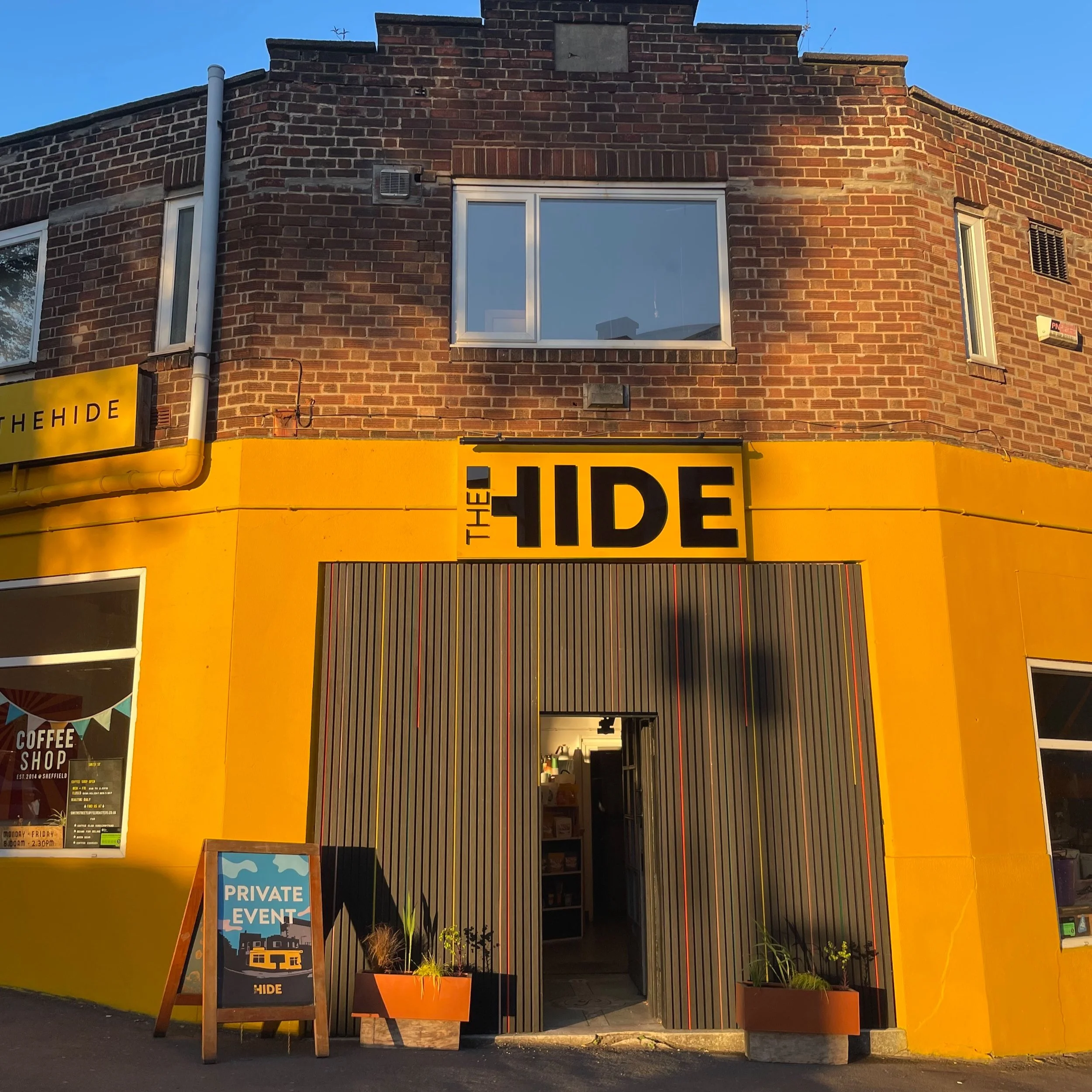 Images shows the yellow 1920s building on The Hide, weddings and events venue in Sheffield. The building is painted a mustard yellow and there is black panelling at the entrance, with copper clouds planters either side of the door.