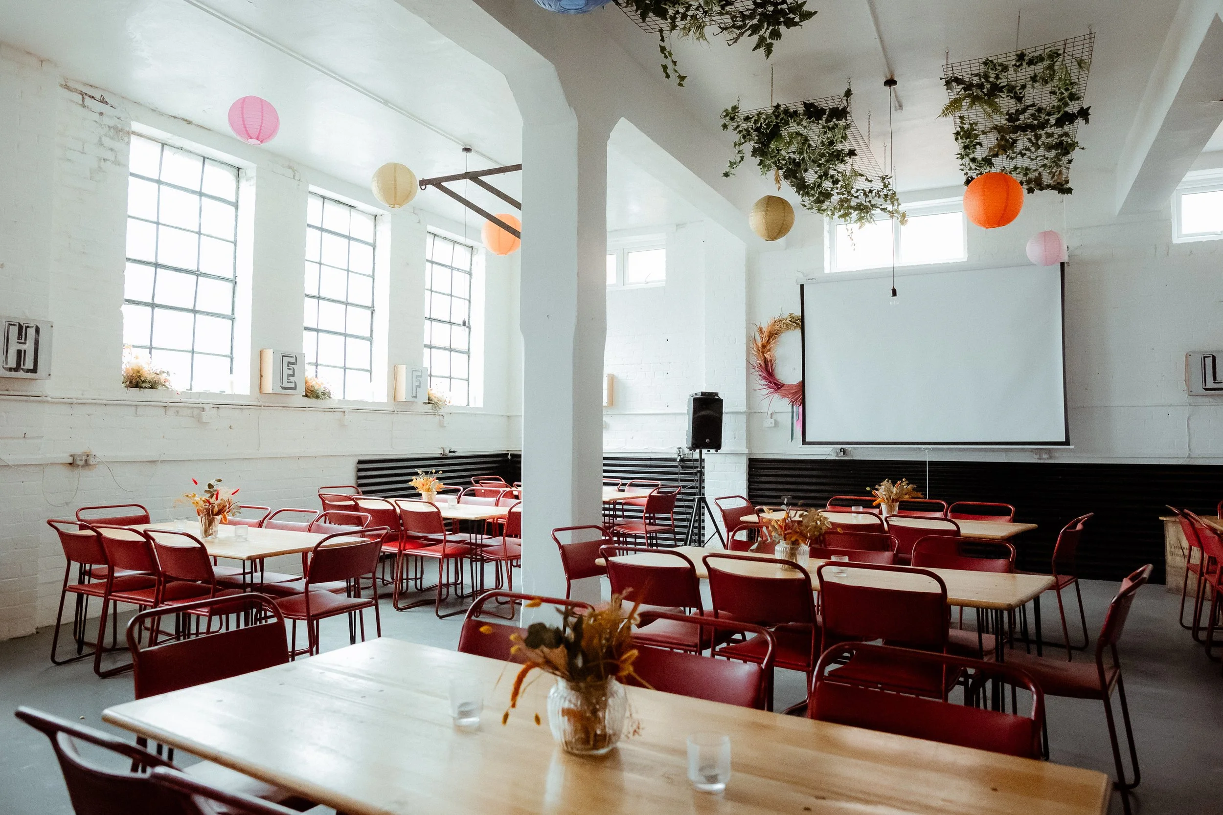 Interior of a decorated event space with high windows, red chairs around wooden tables, hanging paper lanterns, dried floral centerpieces, and a large white projection screen on the wall.
