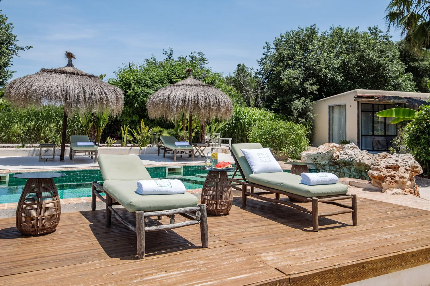 Poolside area with lounge chairs, towels, and a small table with drinks. Two thatched umbrellas and a lush green backdrop with trees and shrubs. Small building to the right, rocks near the edge of the deck, and clear blue sky overhead.