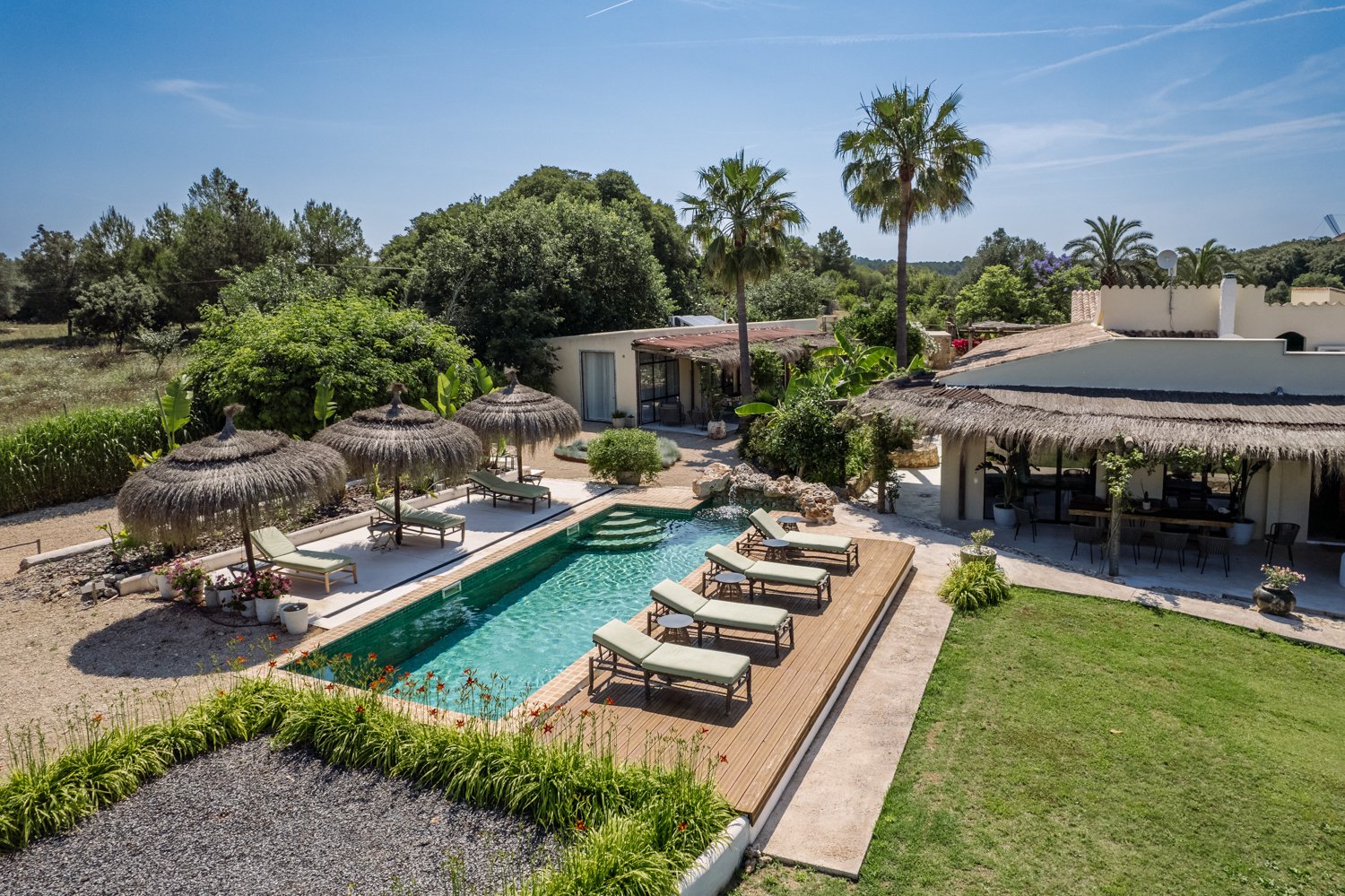 A tropical backyard with a swimming pool, surrounded by lounge chairs with cushions, straw umbrellas, and lush greenery including palm trees, bushes, and potted plants. There is a shaded patio area with outdoor furniture and a building with a thatched roof and white walls in the background under a clear blue sky.