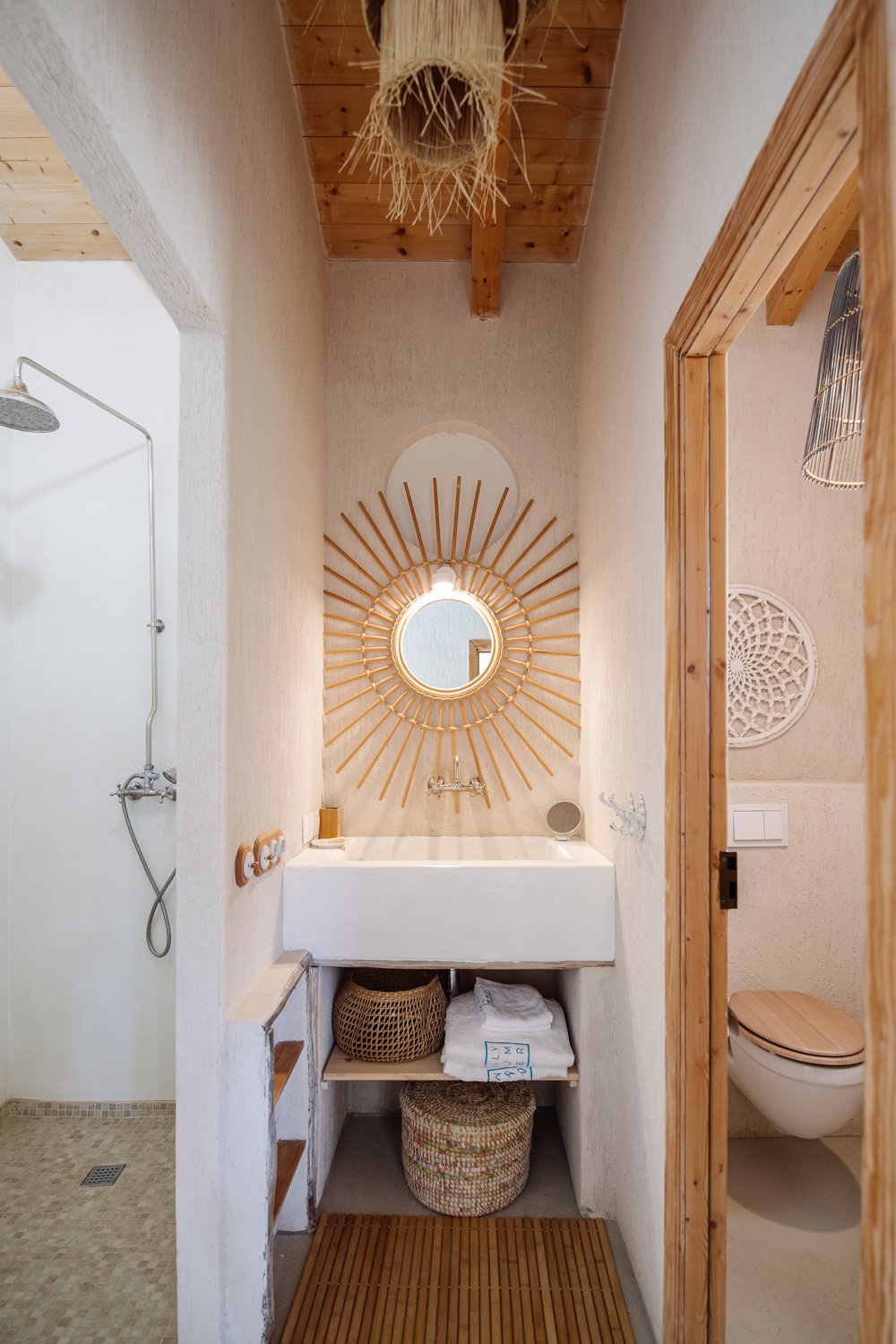 Bathroom with a modern white sink, round mirror with sunburst wooden frame, and wooden accents, including a wooden toilet seat and shelf, with a shower area on the left and a toilet on the right. The decor features natural textures and light colors.