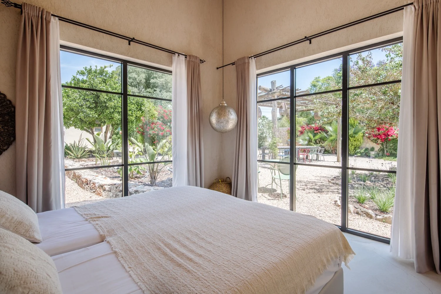 A bedroom with large glass windows showing a outdoor garden with greenery and pink flowers.