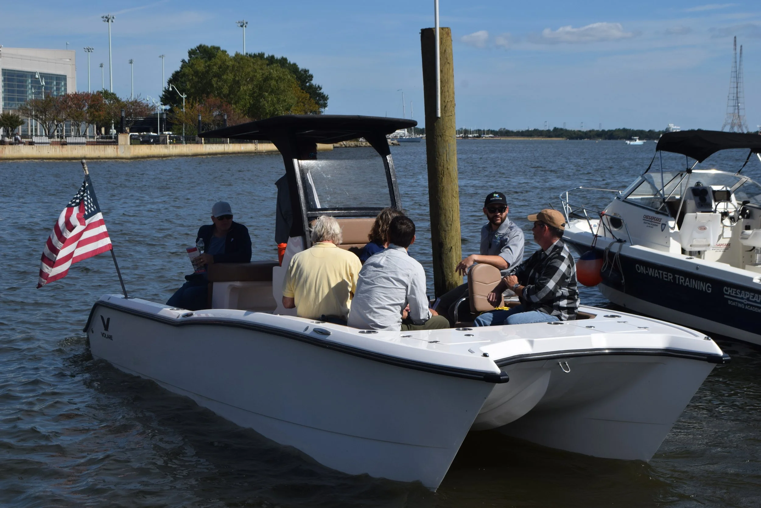 Group of people on a Volare all-electric boat easily holding multiple people and showcasing an American flag, on a body of water with another docked boat nearby, and a cityscape with trees and buildings in the background.