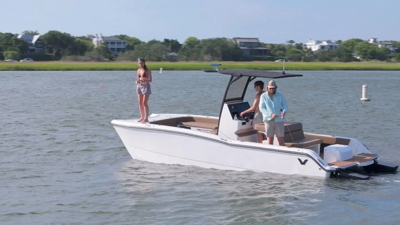 A group of three people on a white motorboat in a calm body of water with houses and greenery in the background.