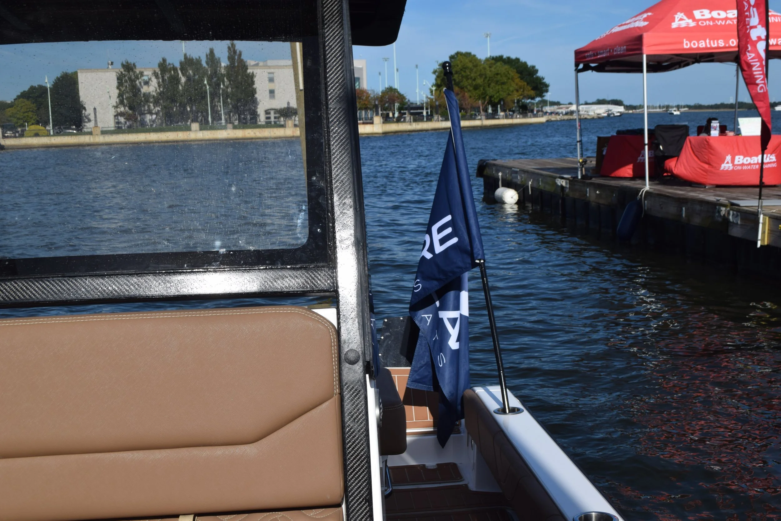 View from a boat dock showing a tan seat, a blue flag of a Volare boat, and a red canopy with tables on a calm waterway, with buildings and trees in the background.