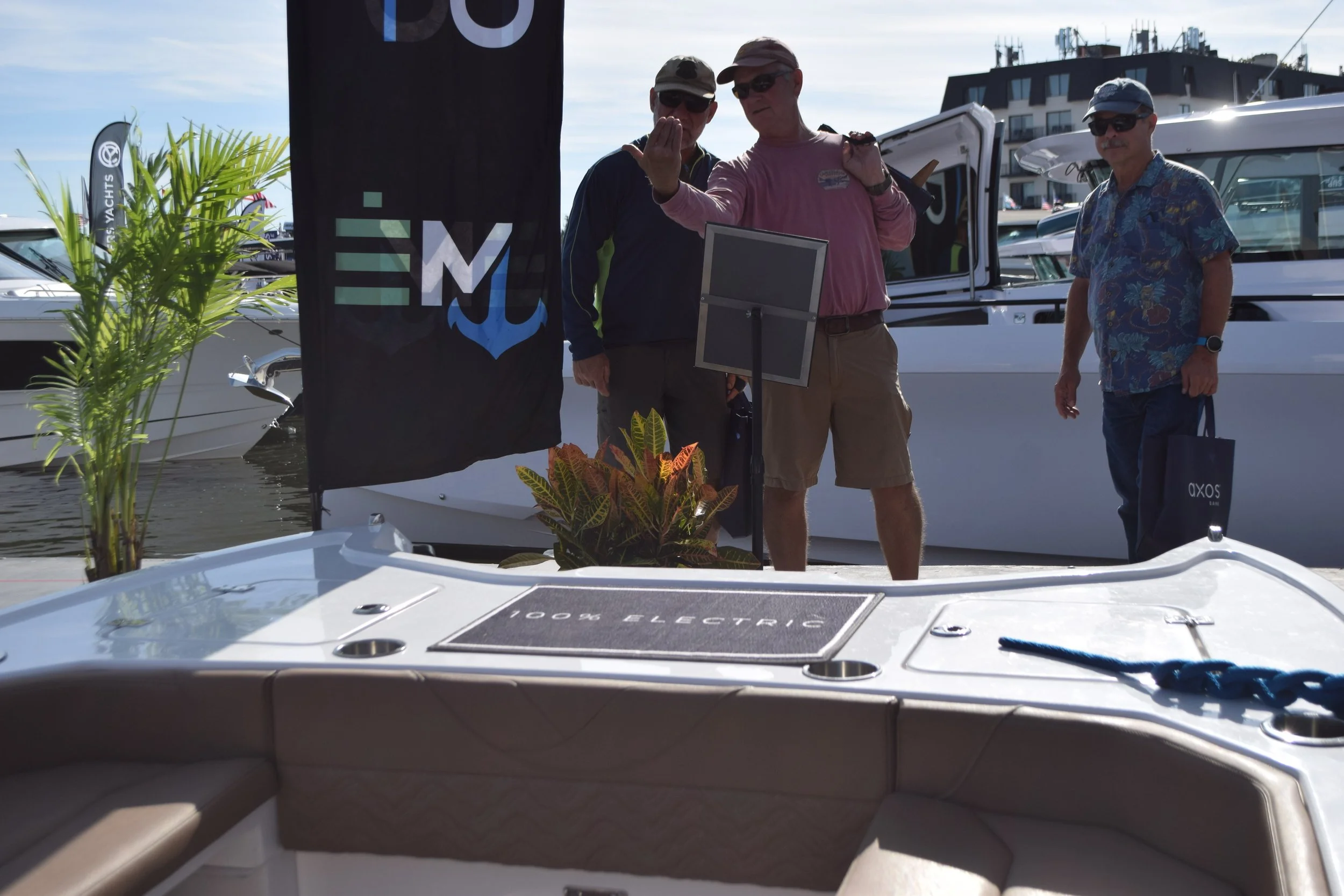 Three men standing on a dock a Volare all-electric boat, one holding a small device, with a black flag showing a blue anchor and the word 'EN' behind them. The foreground shows part of a white electric boat with a sign reading '100% ELECTRIC' on it.