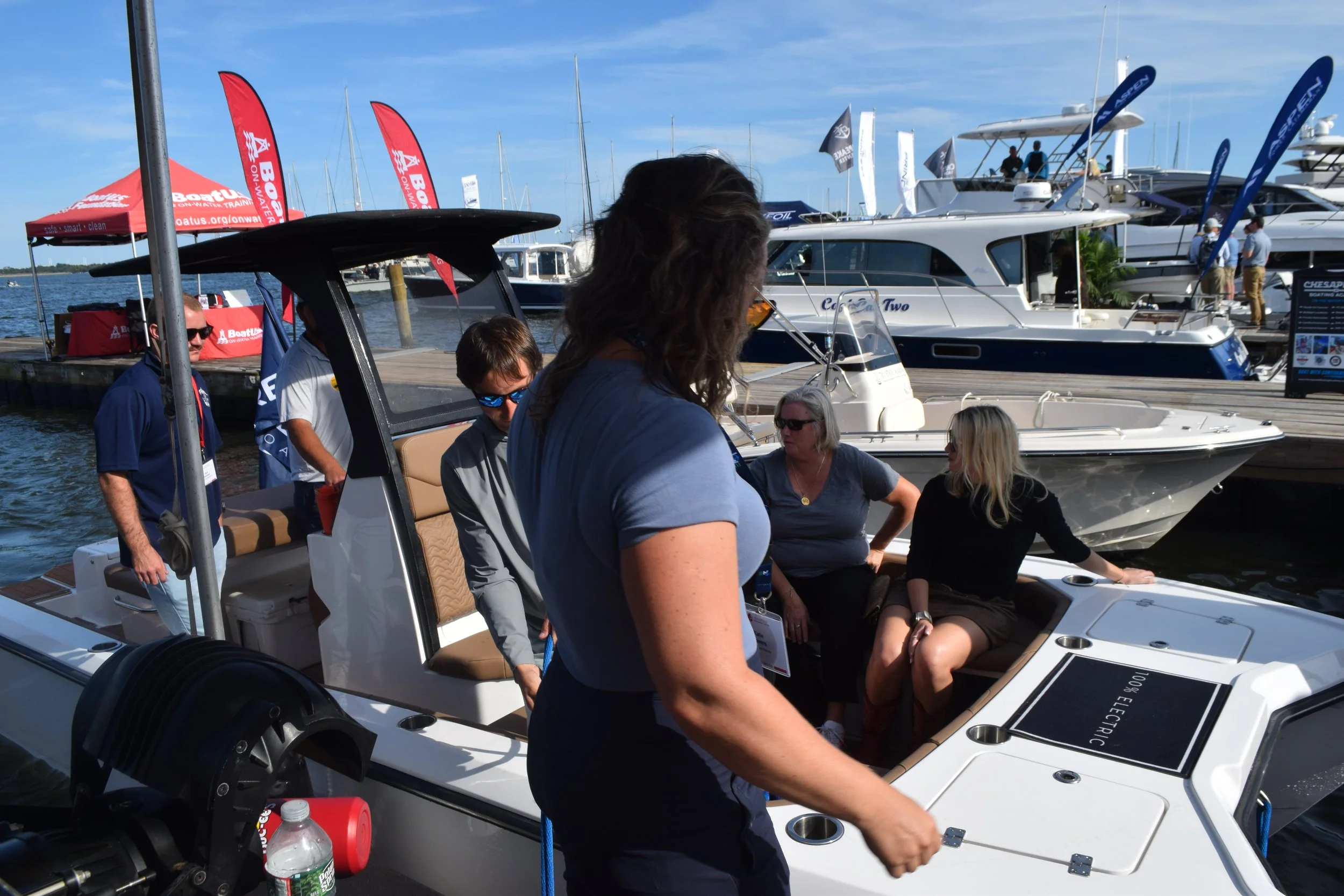 People on a dock inspecting the Volare Boats model Artemis 23 all-electric boat at a busy boat show, with yachts and banners in the background.
