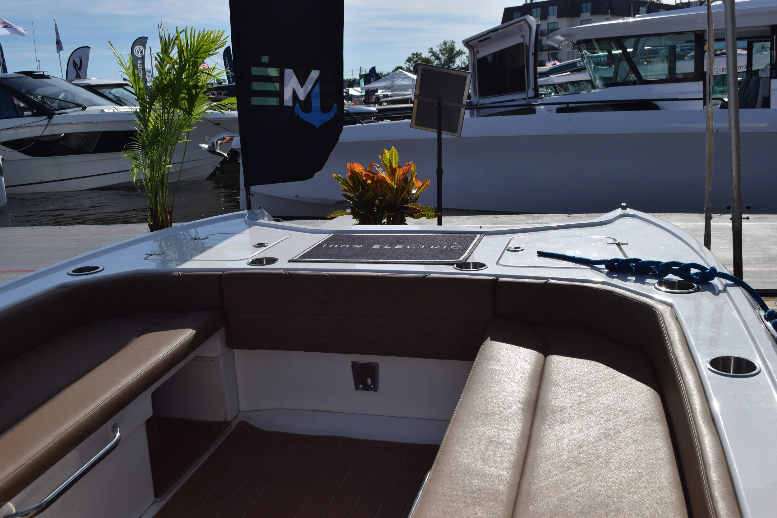 View of a Volare boat's interior with sleek, luxurious beige cushioned seating, a small potted plant, and a sign reading '100% Electric,' with boats and marina in the background.