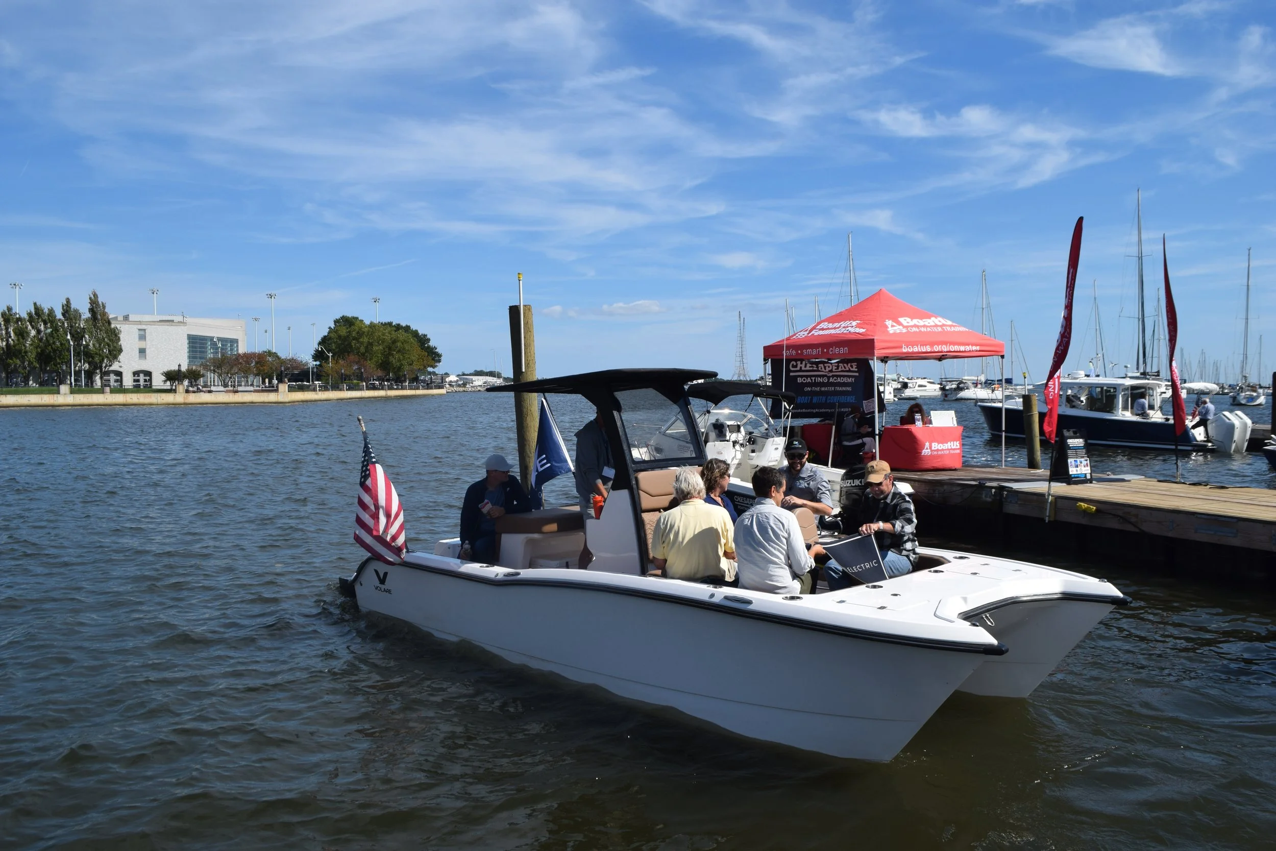 People riding on Volare Boats' flagship Artemis 23 all-electric boat near a pier with sailing boats and a red canopy in the background against a blue sky.