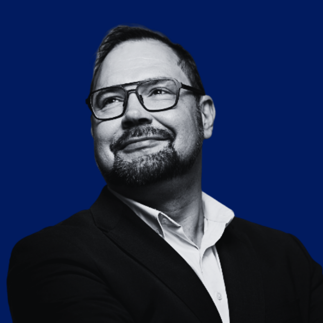 Black and white portrait of a man with glasses, short hair, and a beard, smiling and looking upwards, against a dark blue background.