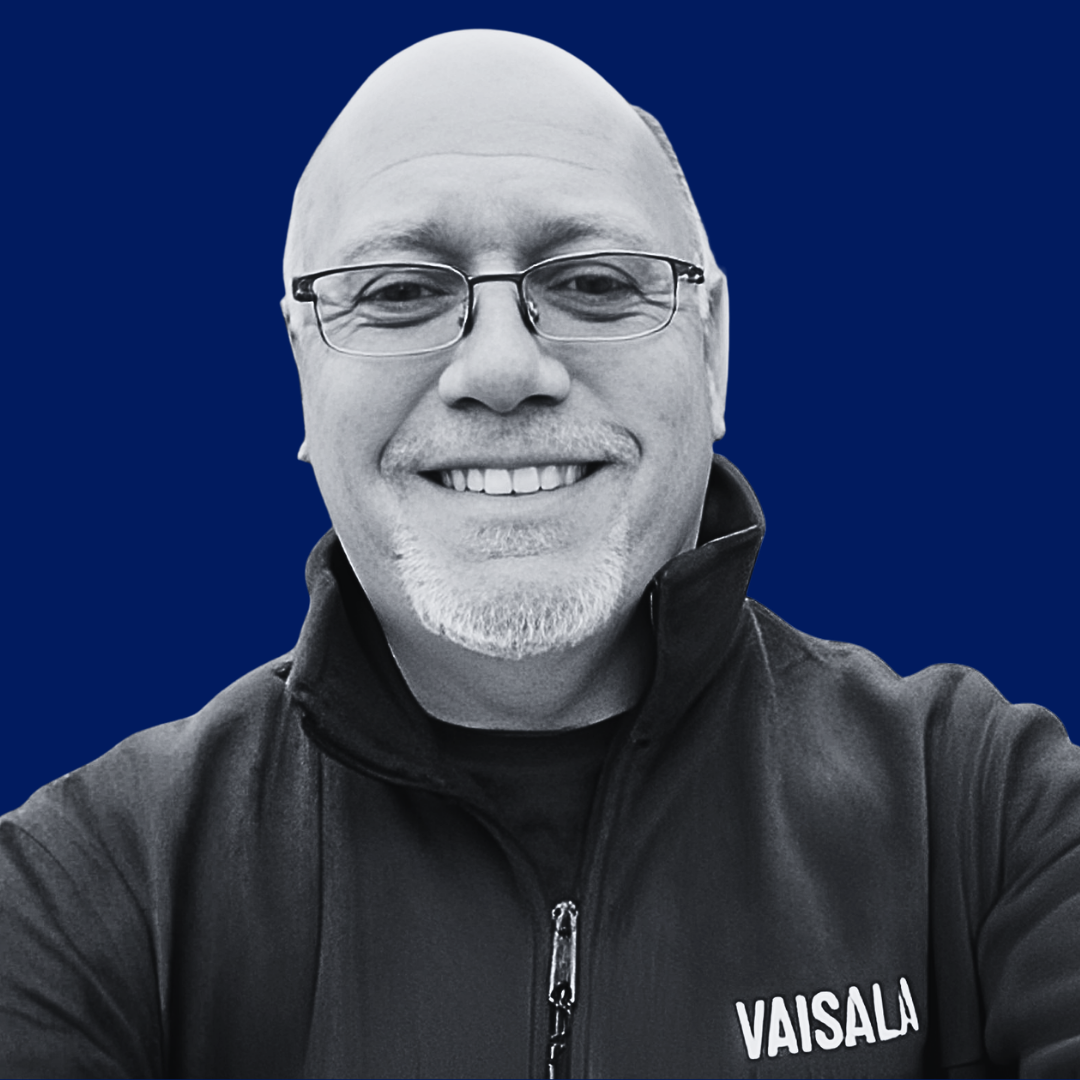 A smiling man with glasses, a goatee, wearing a black jacket with a zipper and the name 'VAISALA' on it, taking a selfie against a dark blue background.