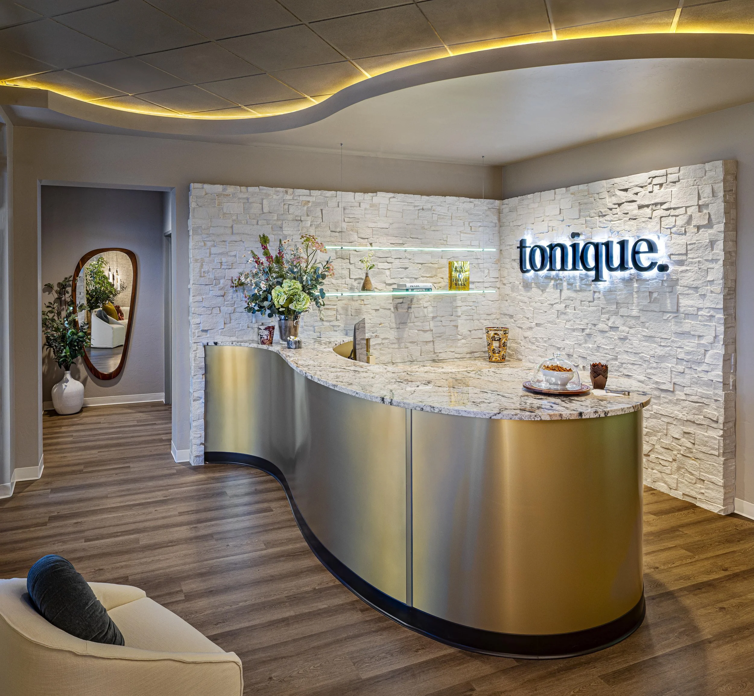 Modern reception area with a curved marble countertop, white textured brick wall, illuminated 'tonique' sign, and decorative vases with flowers and books.