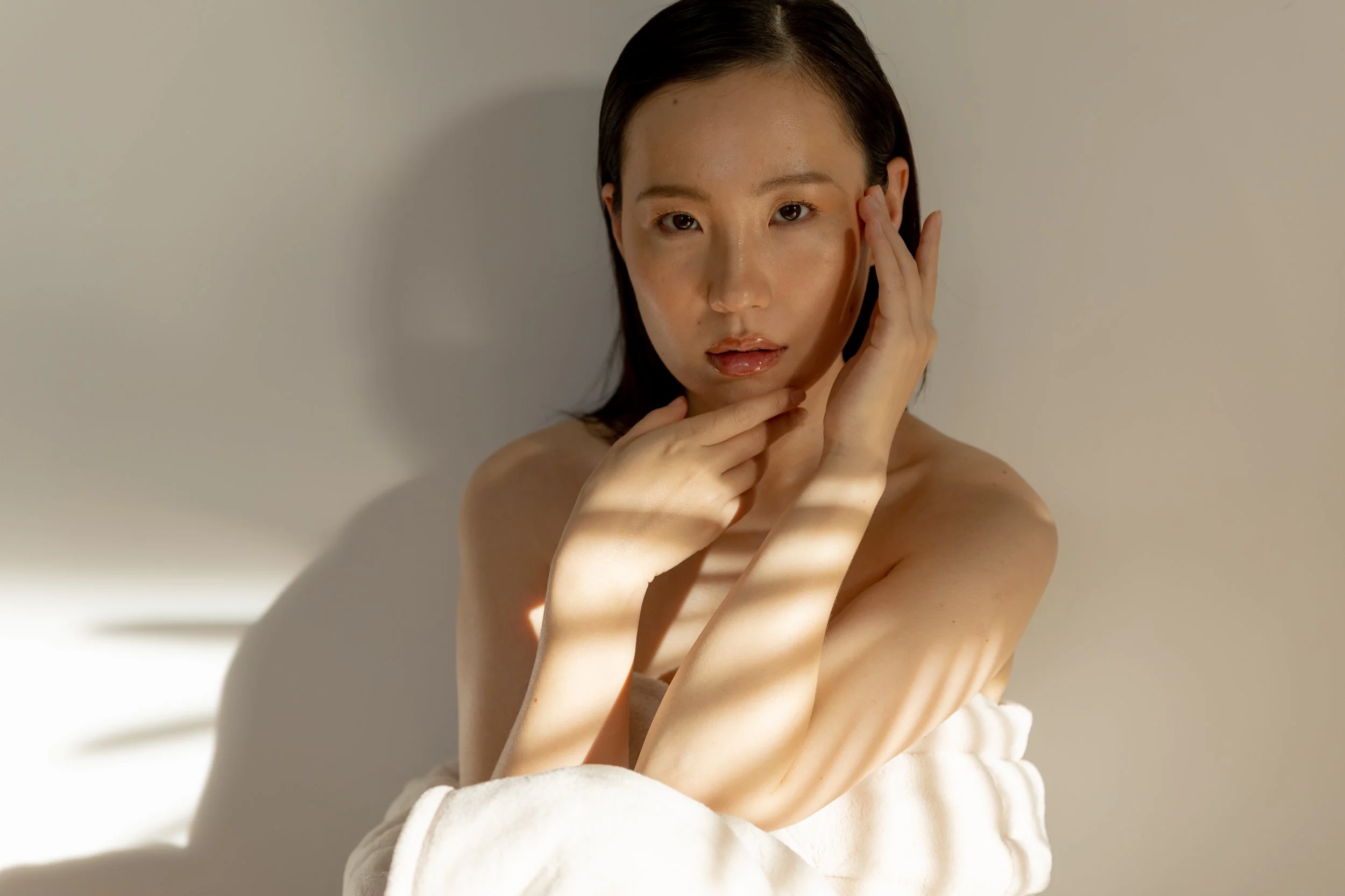 A young woman with dark hair poses against a plain wall, partially wrapped in a white towel, with light casting striped shadows across her face and body.