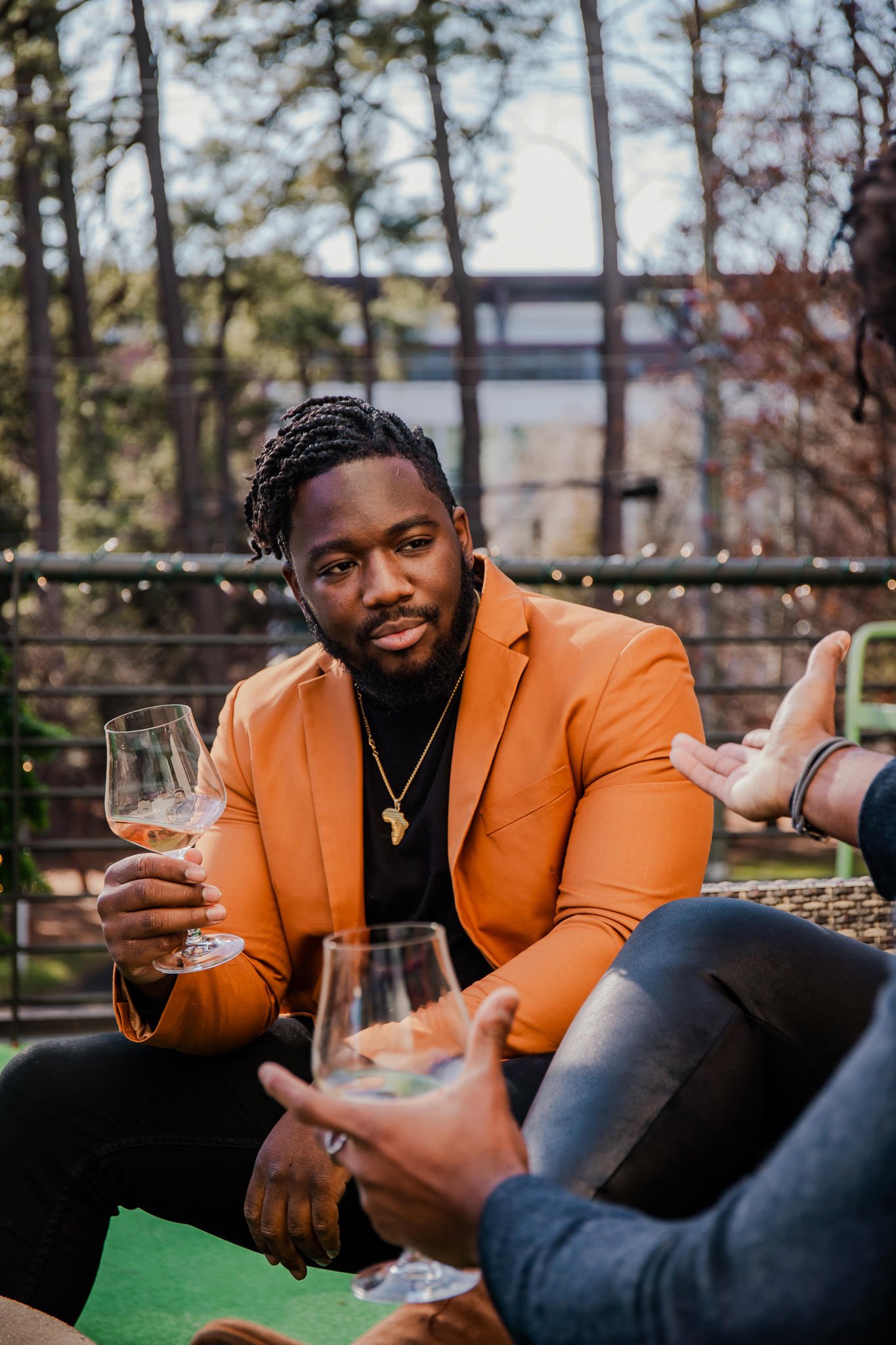 Two people having a conversation outdoors, one holding a wine glass, wearing an orange blazer.