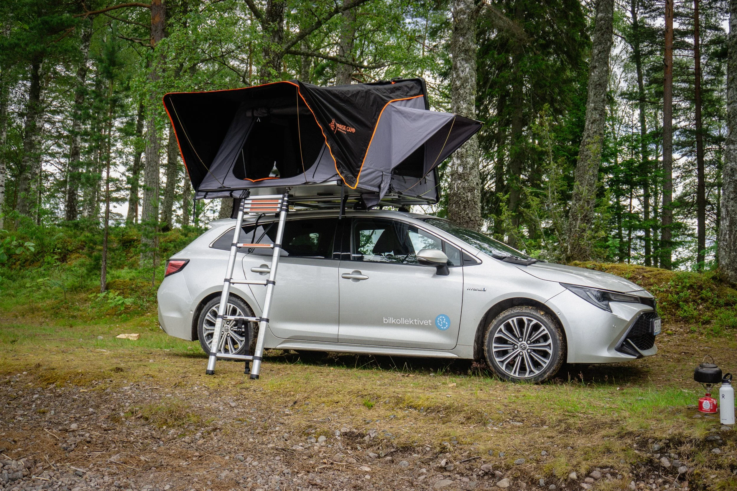En sølvfarget bil med takboks installert på taket, parkert i en skog med trær og røtter. Det er en trappestige festet til bilen for å komme opp til takboksen. Det står logo og tekst på siden av bilen som sier «bilkollektivet».