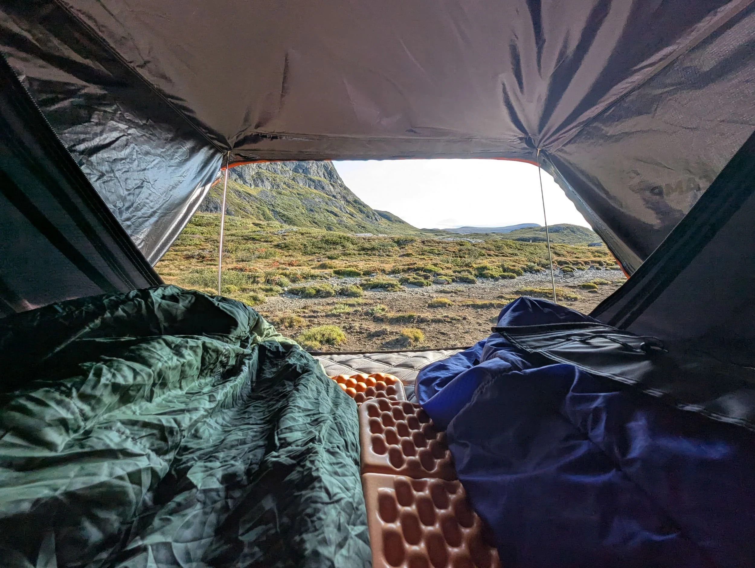 Utsikt fra innsiden telt med åpent dør, viser et fjellandskap med grønne åser og en blå himmel i bakgrunnen.