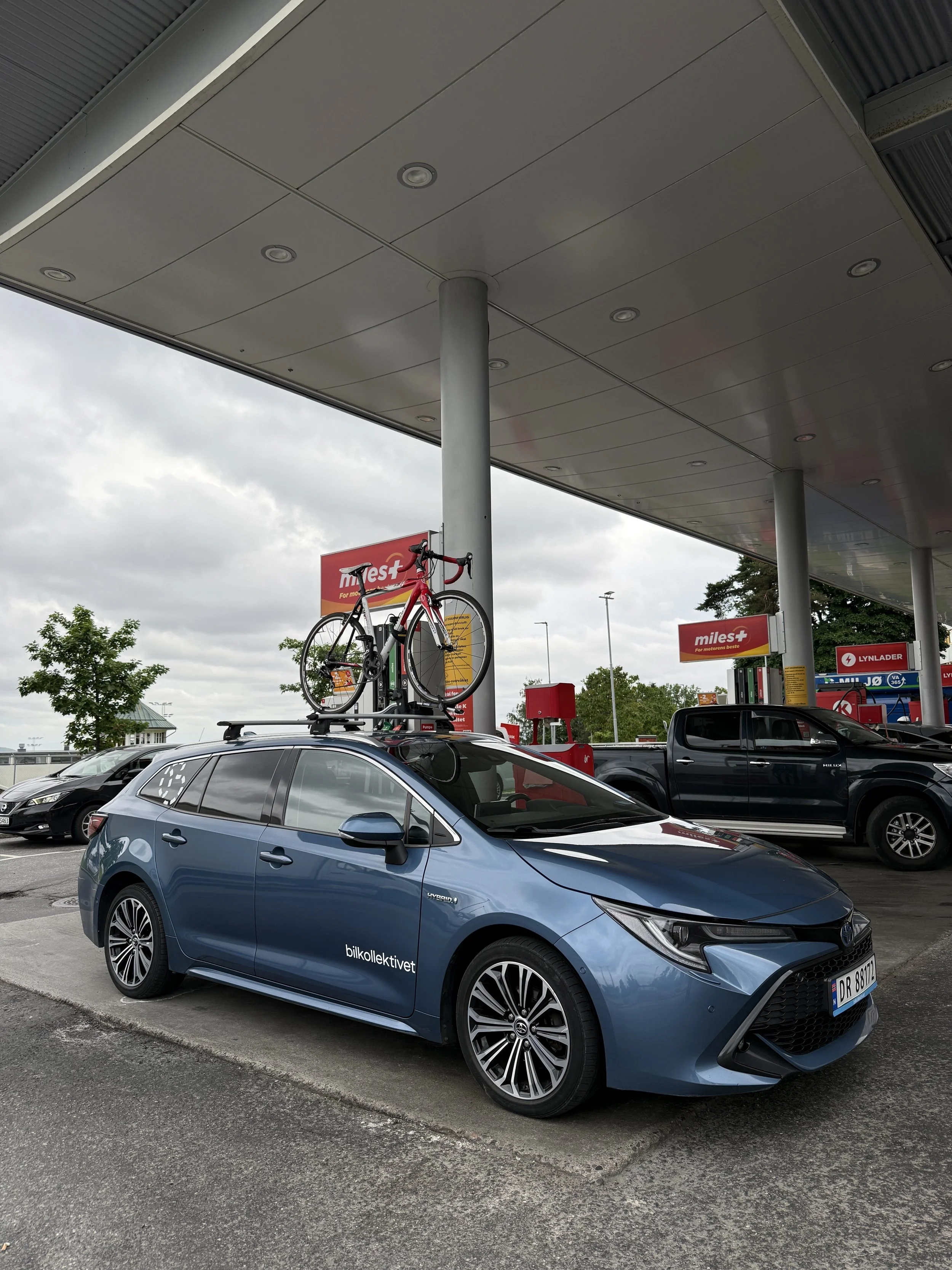 Bilde av en bensinstasjon med flere biler parkert. En av bilene har en sykkel montert på taket.