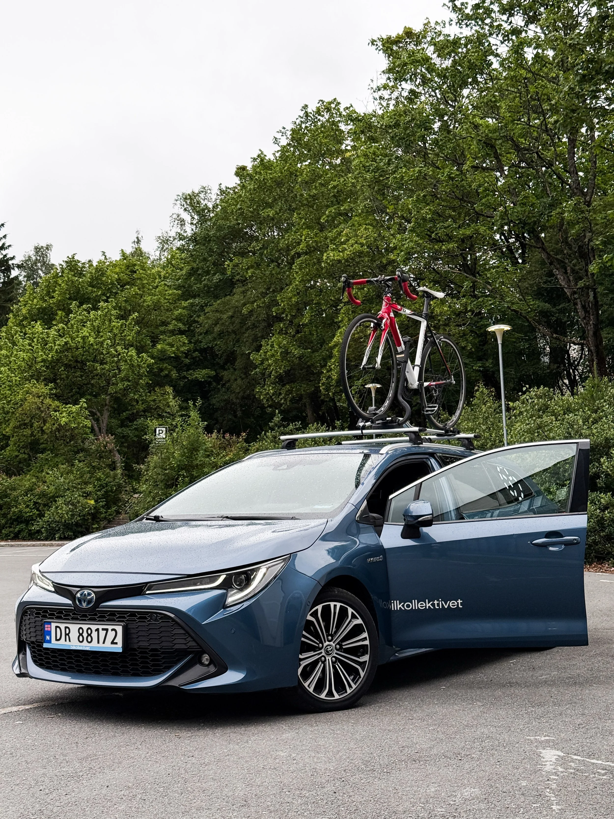 En blå Toyota hybridbil med registreringsnummer DR 88172, med en sykkeleie på taket, parkert i en parkeringsplass mot en bakgrunn av grønne trær og overlys.