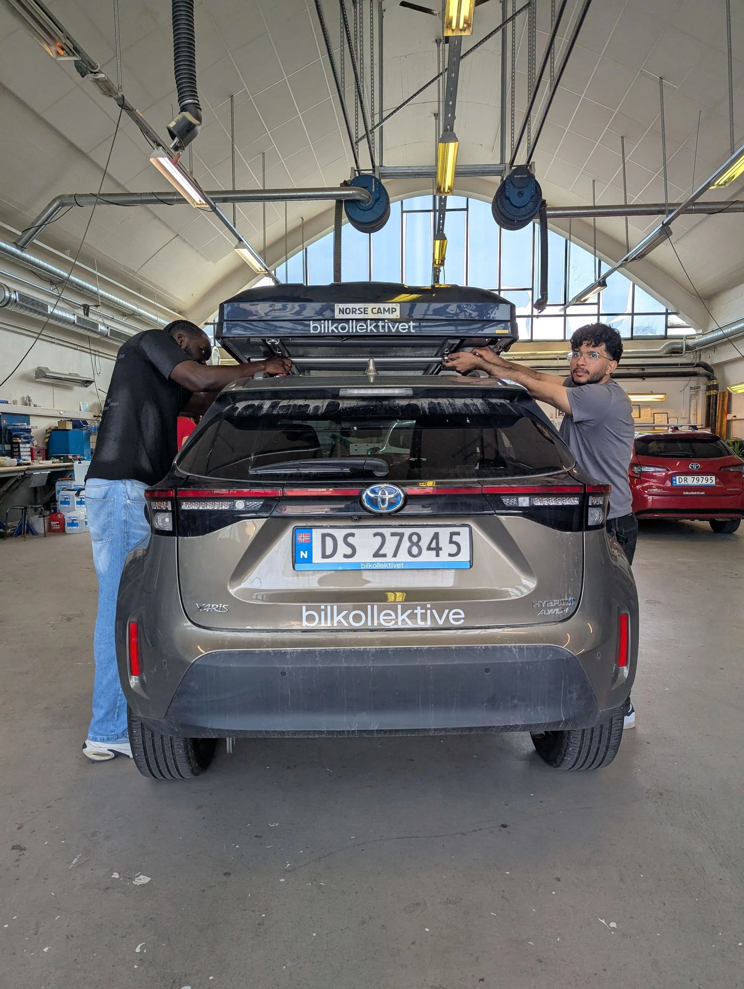 To personer jobber på taktelt av en svart Toyota Yaris cross . De er i et verksted med verktøy og utstyr på veggen, og det er store vinduer i taket som slipper inn naturlig lys.