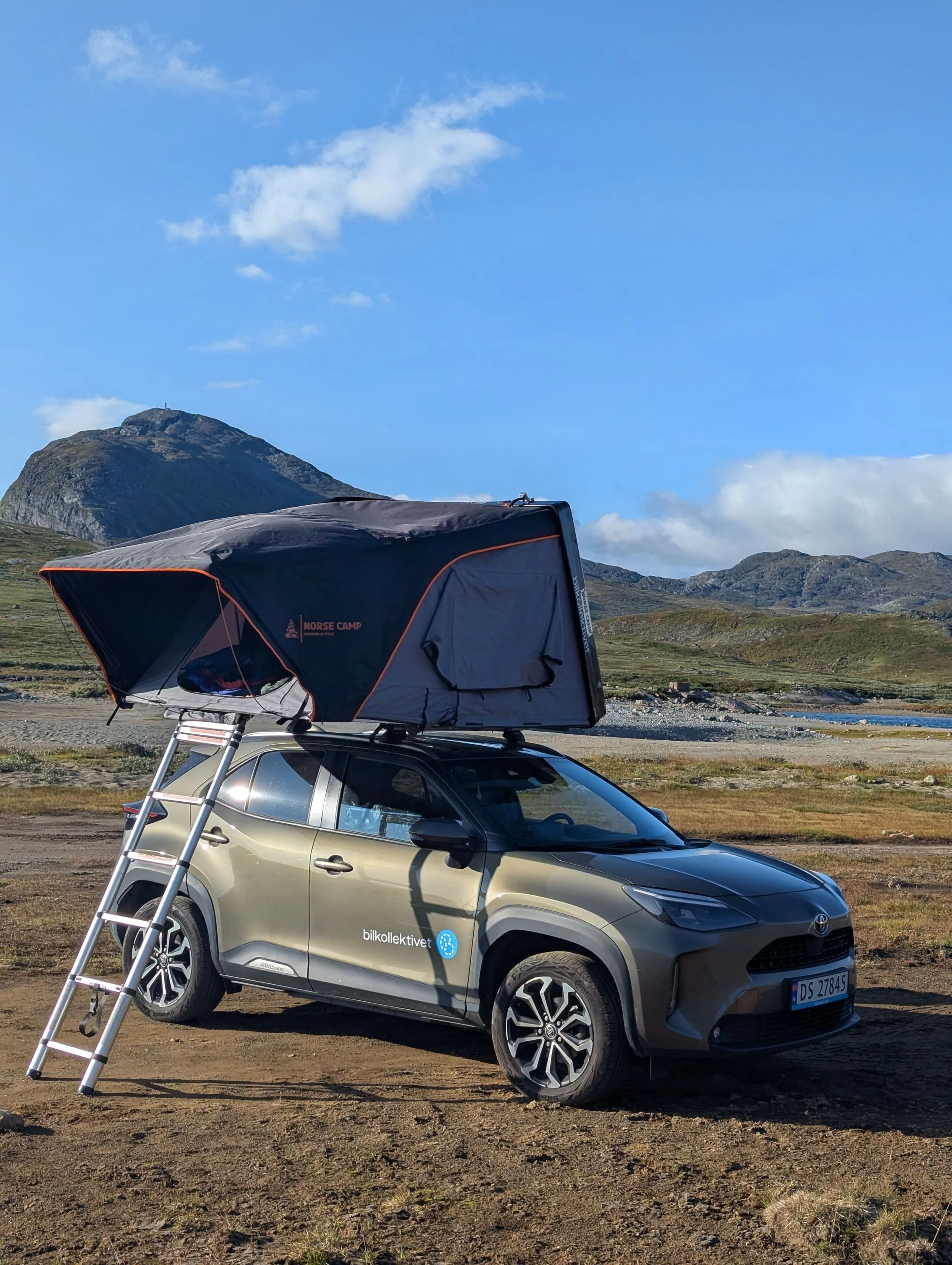 En grå bil med taktelt på taket, parkert på en jordflekk ved fjell med blå himmel og noen skyer. Takkuppelen er fra Norse Camp.